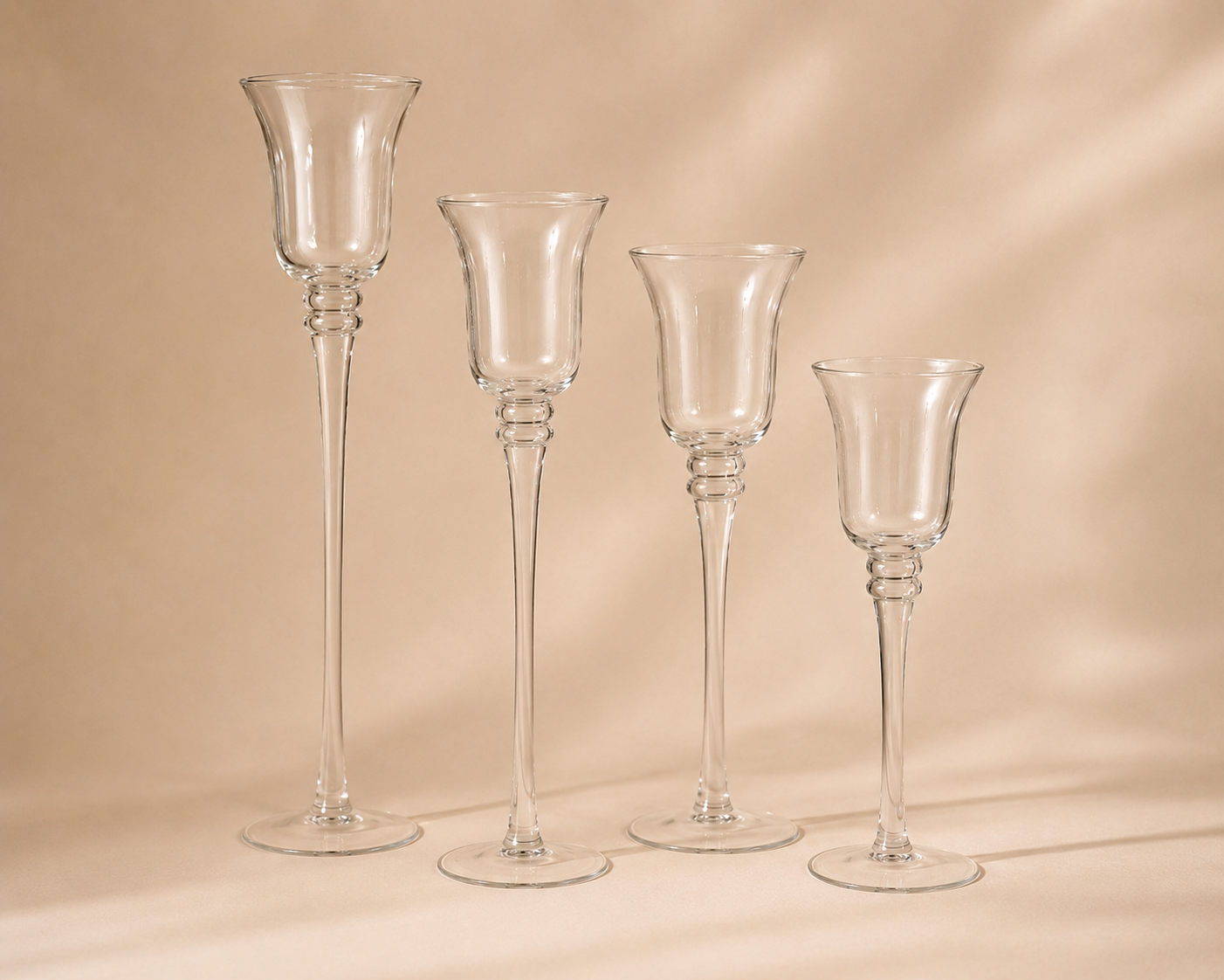 Four tall, clear glass vases with long, slender stems and flared openings, arranged in a row against a plain beige background.