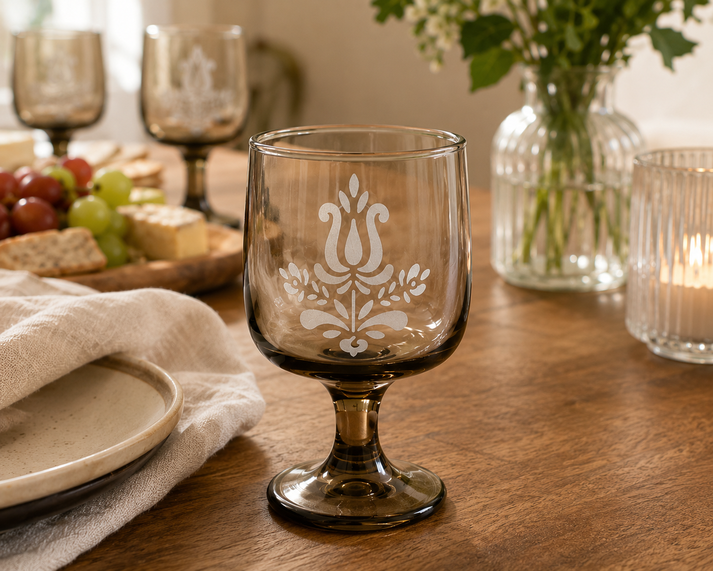 A decorative glass with a white floral and leaf pattern etched on it, placed on a wooden dining table with a background of candles, a vase of green leaves, grapes, cheese, and other glassware.