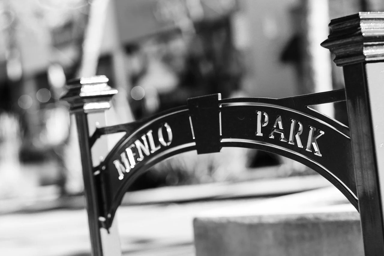 Black and white photo of a park sign with the words "MENLO PARK" on a curved metal plaque, with blurred background.