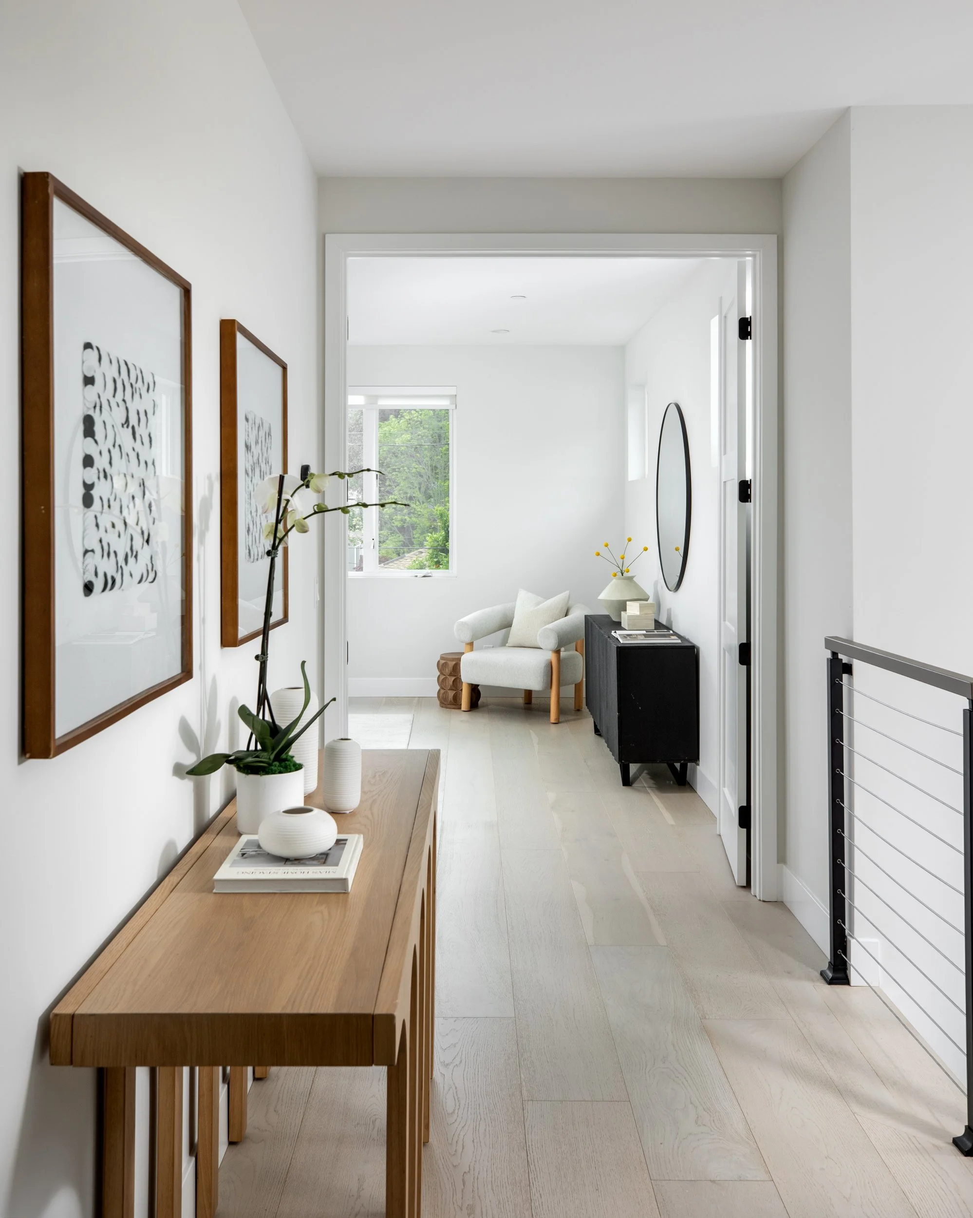 A hallway with light wooden flooring and white walls, decorated with framed abstract black and white art, leading to a sitting area with a white armchair, black sideboard, round mirror, and a window showing greenery outside.