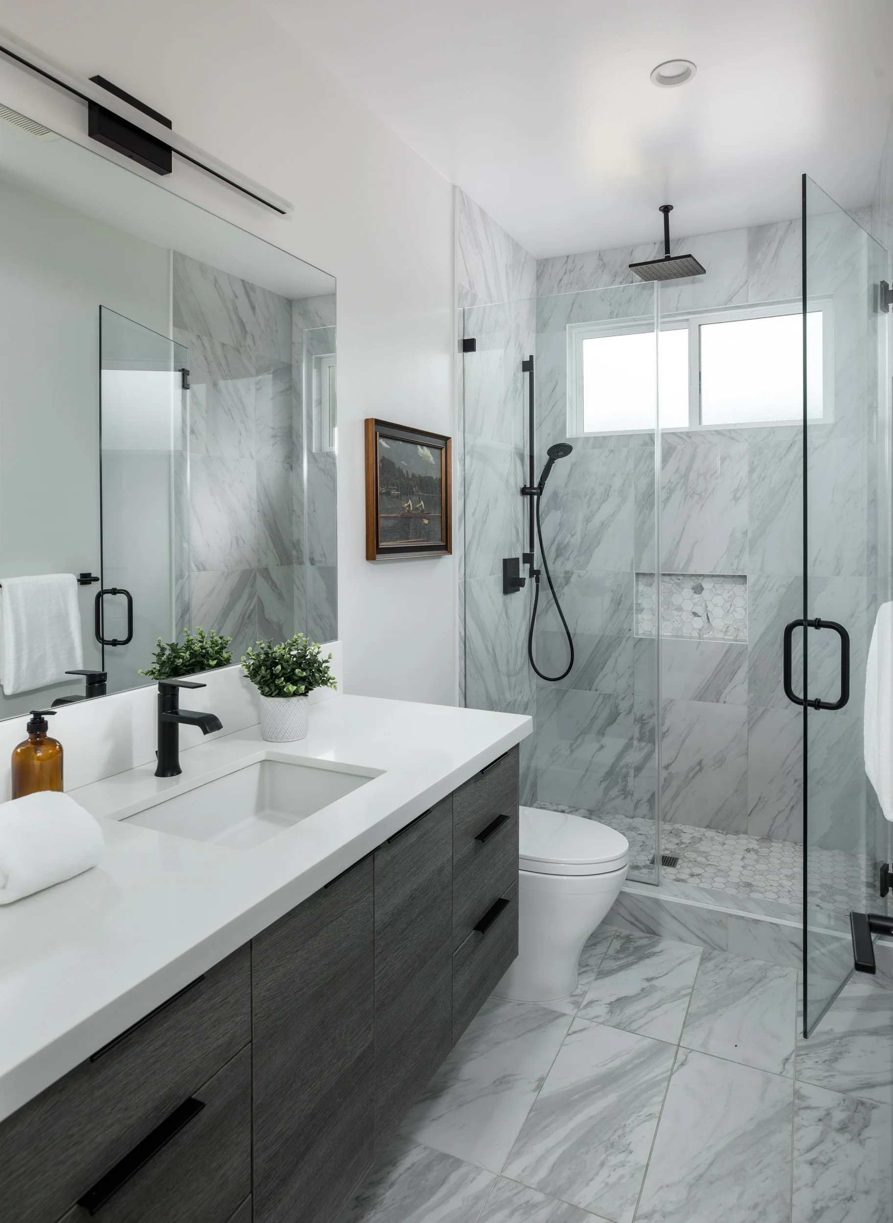 Modern bathroom with marble tile walls and floors, a glass shower with black fixtures, a dark wood vanity with a white countertop, bedside plants, a framed art piece, and a large mirror.