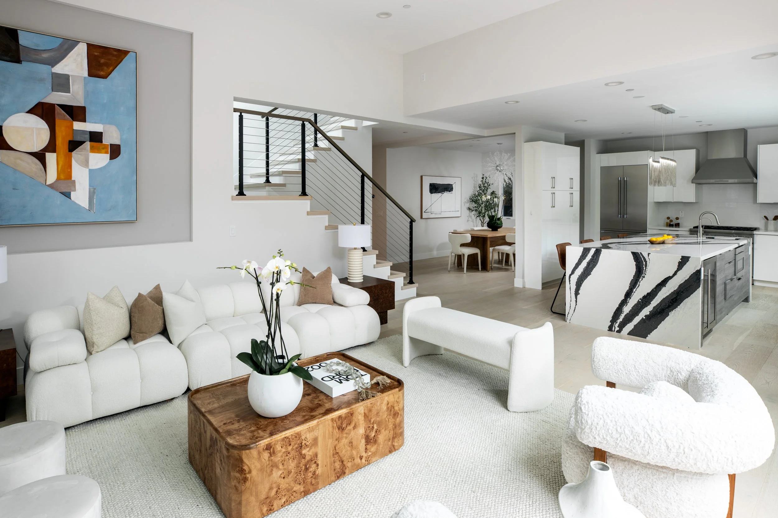 Modern open-concept living room with white sofa, wooden coffee table with orchid, black and white abstract painting, staircase with black railing, dining area with wooden table and chairs, kitchen with marble island, stainless steel appliances, and w