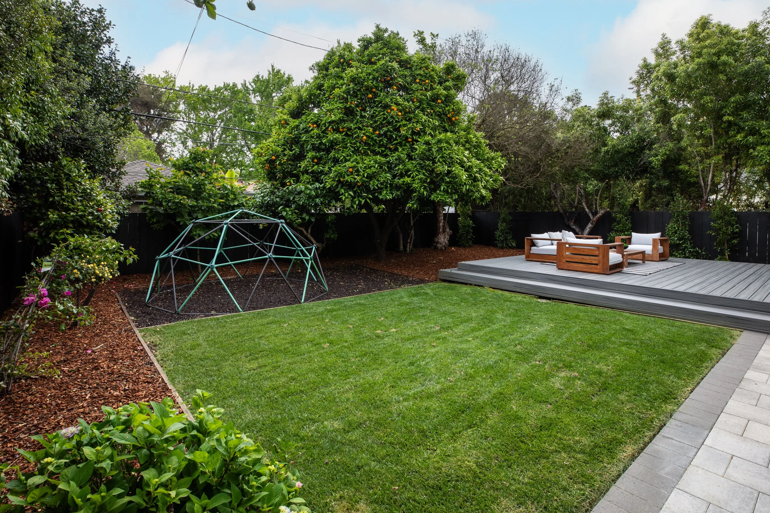 A backyard outdoor space featuring a grassy lawn, a small play structure, a wooden deck with outdoor furniture, and lush trees and shrubs.