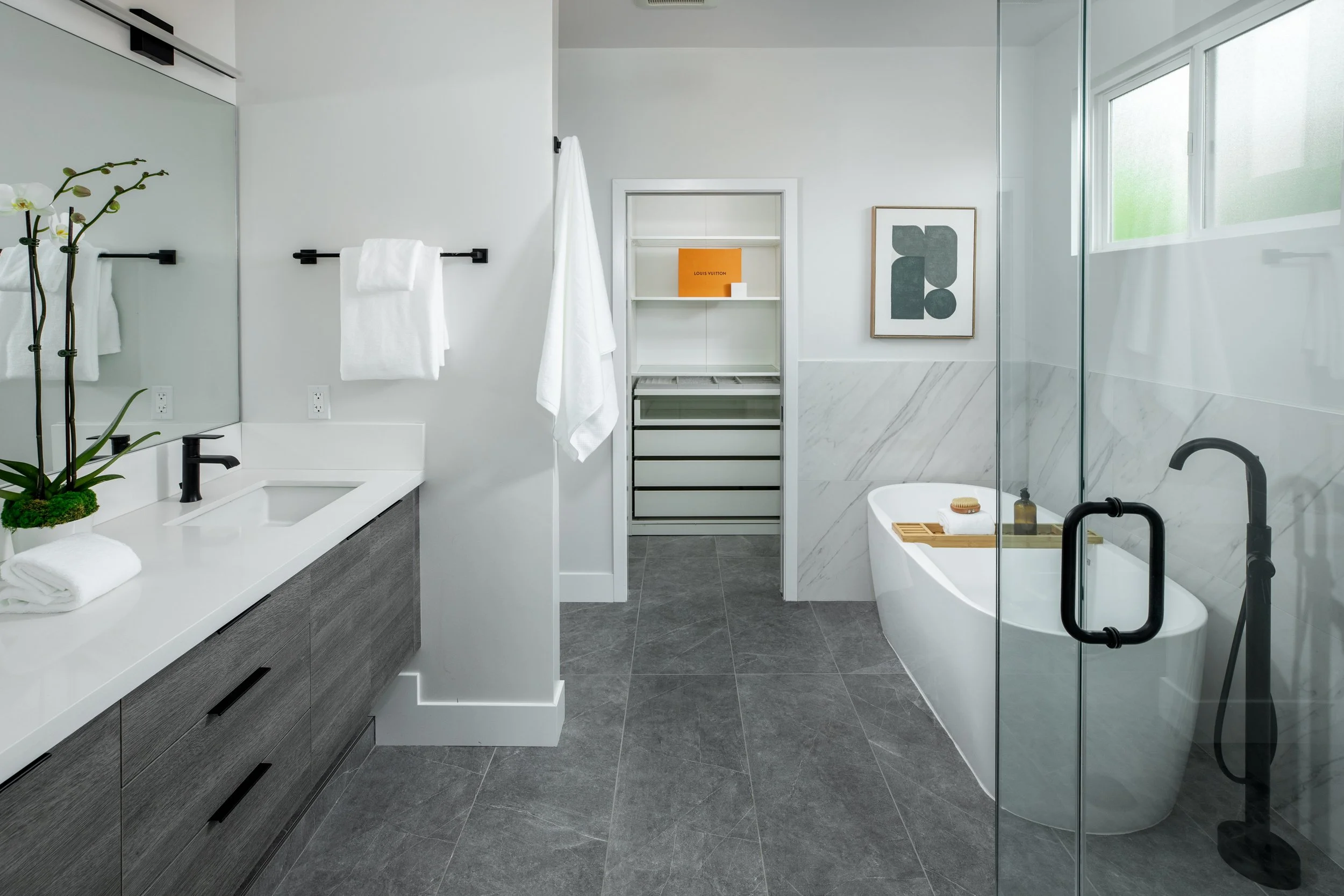 Modern bathroom featuring a white bathtub with wooden tray, black fixtures, glass shower enclosure, gray tiled floor, white walls with marble accents, potted orchid, white sink with black faucet, towels on black racks, framed artwork, and a window.