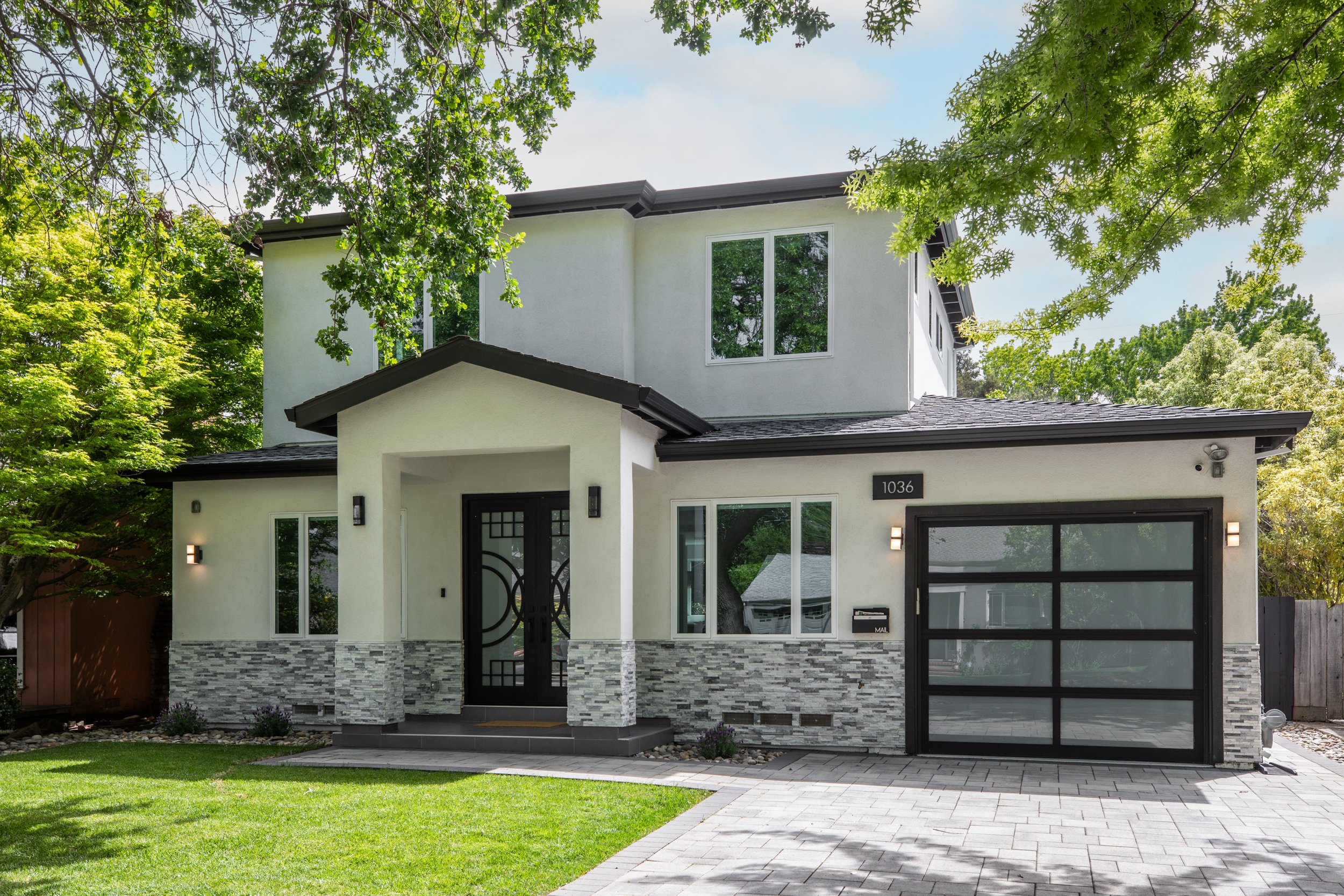 Modern two-story house with white exterior, black trim, and a gray stone accent on the lower walls, surrounded by lush green trees and a well-manicured lawn.