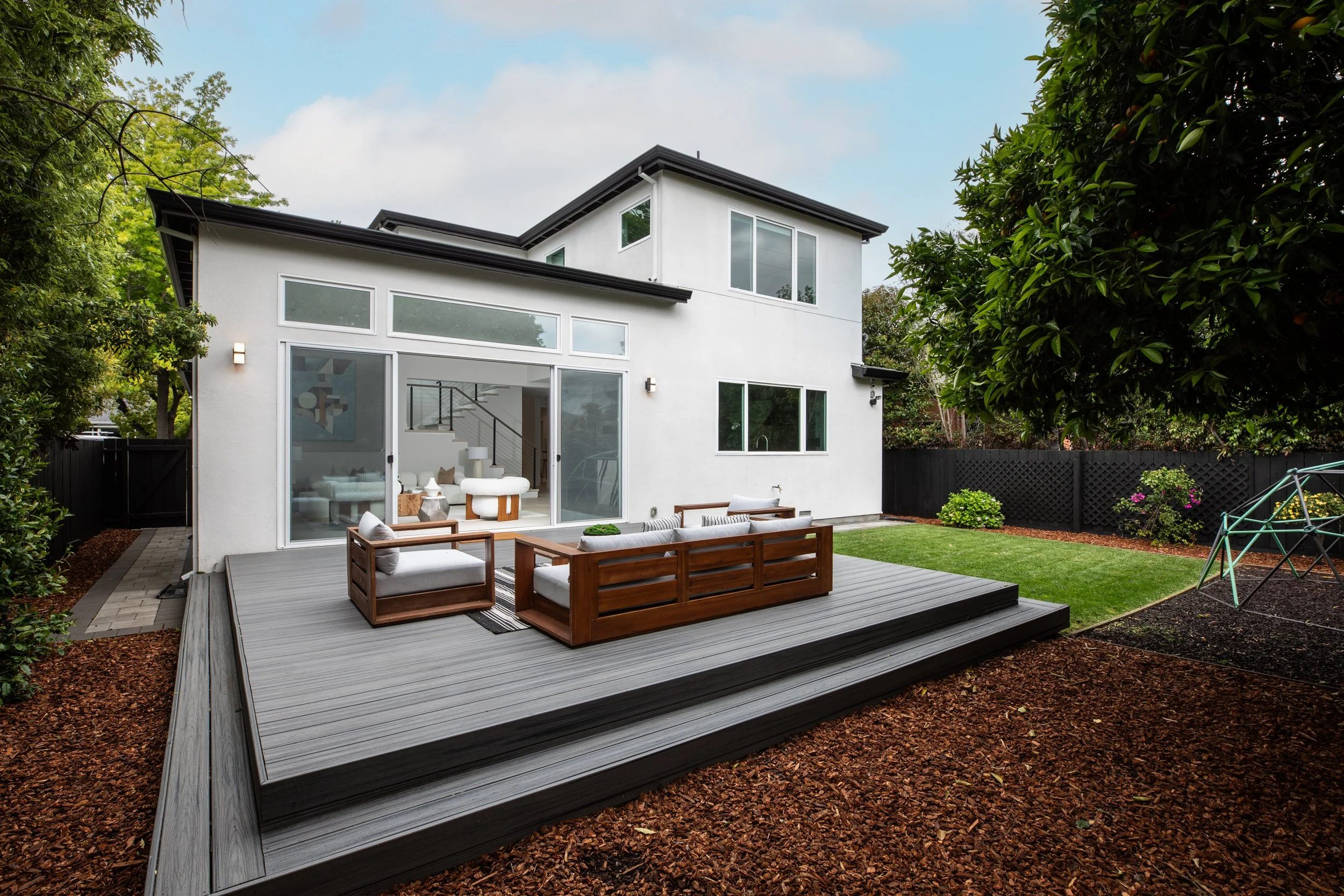 Modern white two-story house with large glass doors opening to a backyard deck with outdoor seating, surrounded by a manicured lawn, trees, and landscaping.