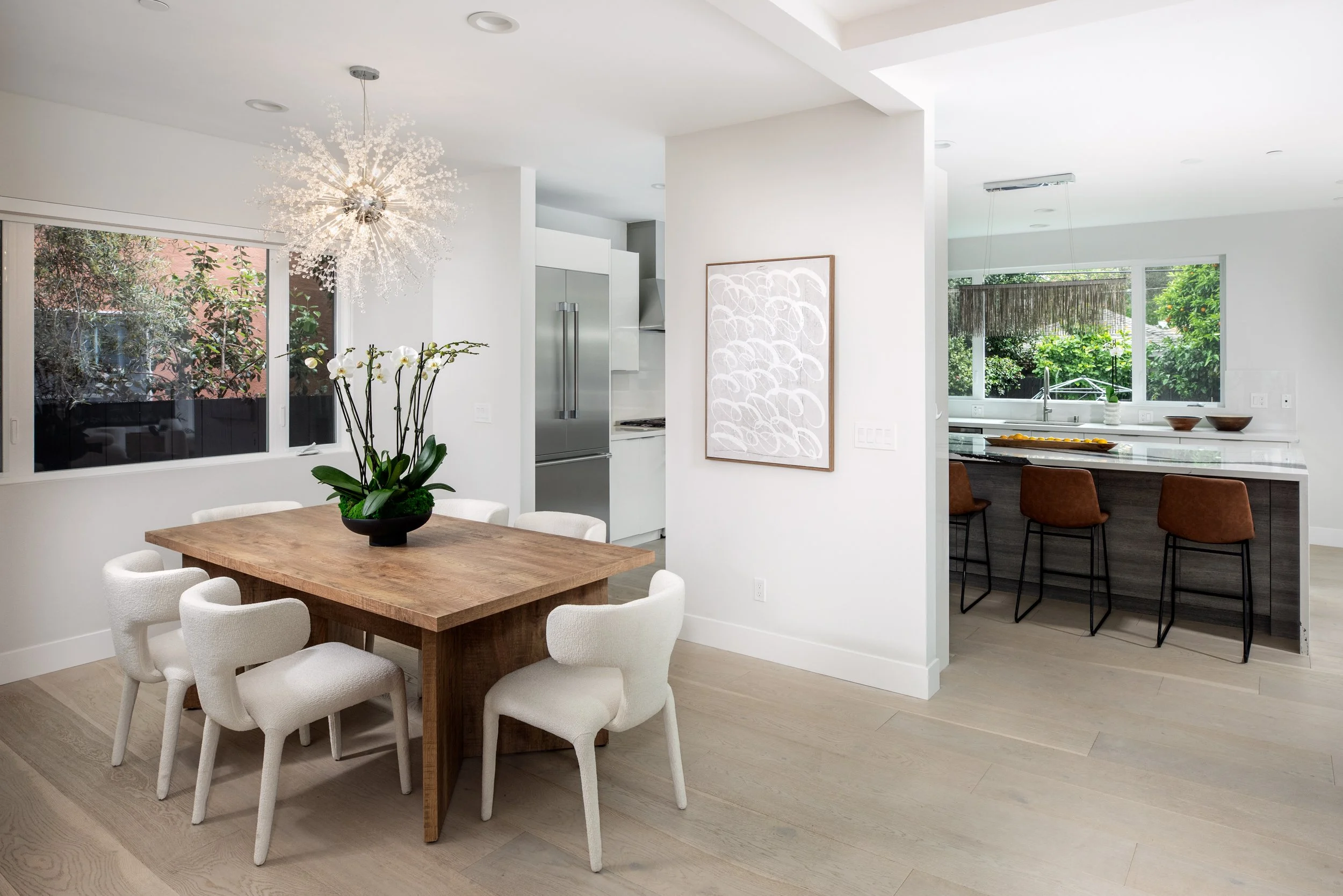Modern dining area with wooden table, white chairs, orchid plant, and chandelier, open to a kitchen with bar stools and large windows.