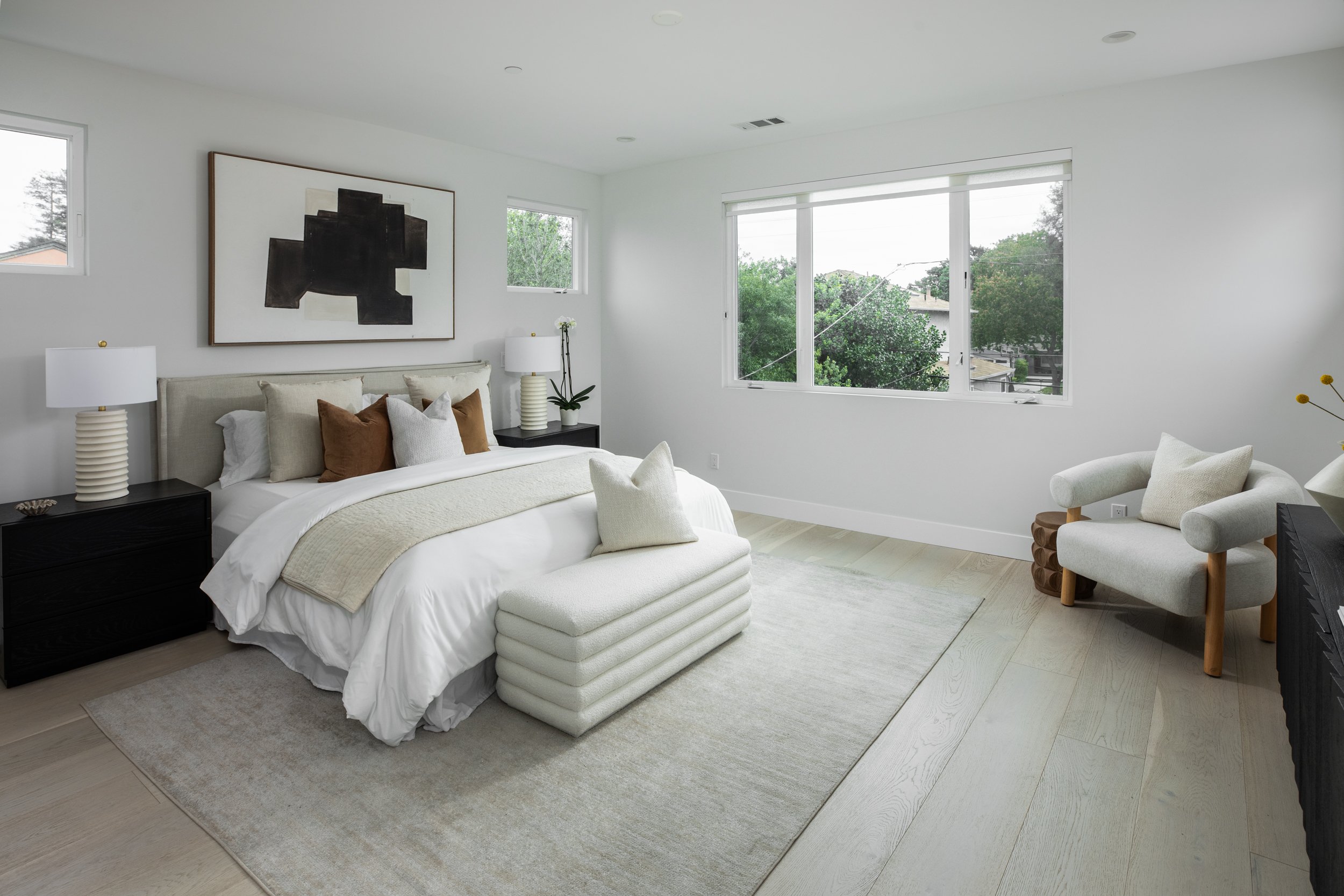 A modern bedroom with white walls, large windows, a bed with white bedding and beige and brown pillows, black nightstands with table lamps, a beige armchair with a pillow, a white ottoman at the foot of the bed, and minimalistic decor.