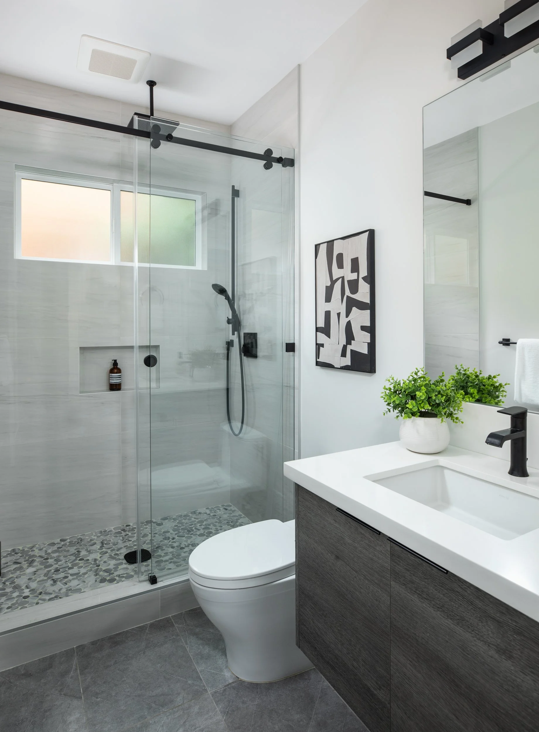 Modern bathroom with glass shower enclosure, small window, gray and white color scheme, black fixtures, and a decorative black and white wall art. Contains a white toilet, a gray vanity with a white sink and a black faucet, and a potted green plant.