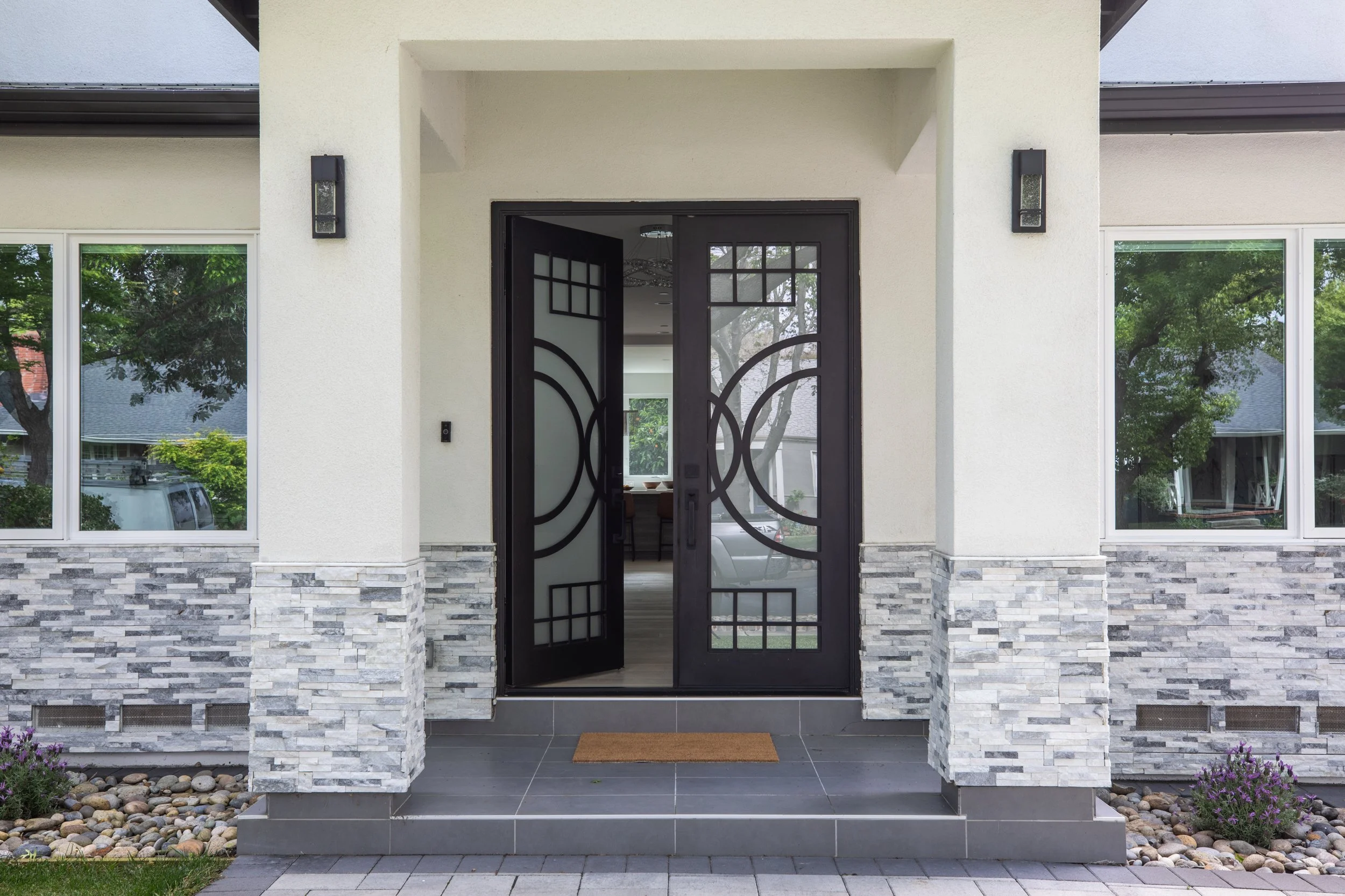 Front door of a modern house with decorative black iron framework, surrounded by white walls, stone accents, and large windows revealing greenery outside.