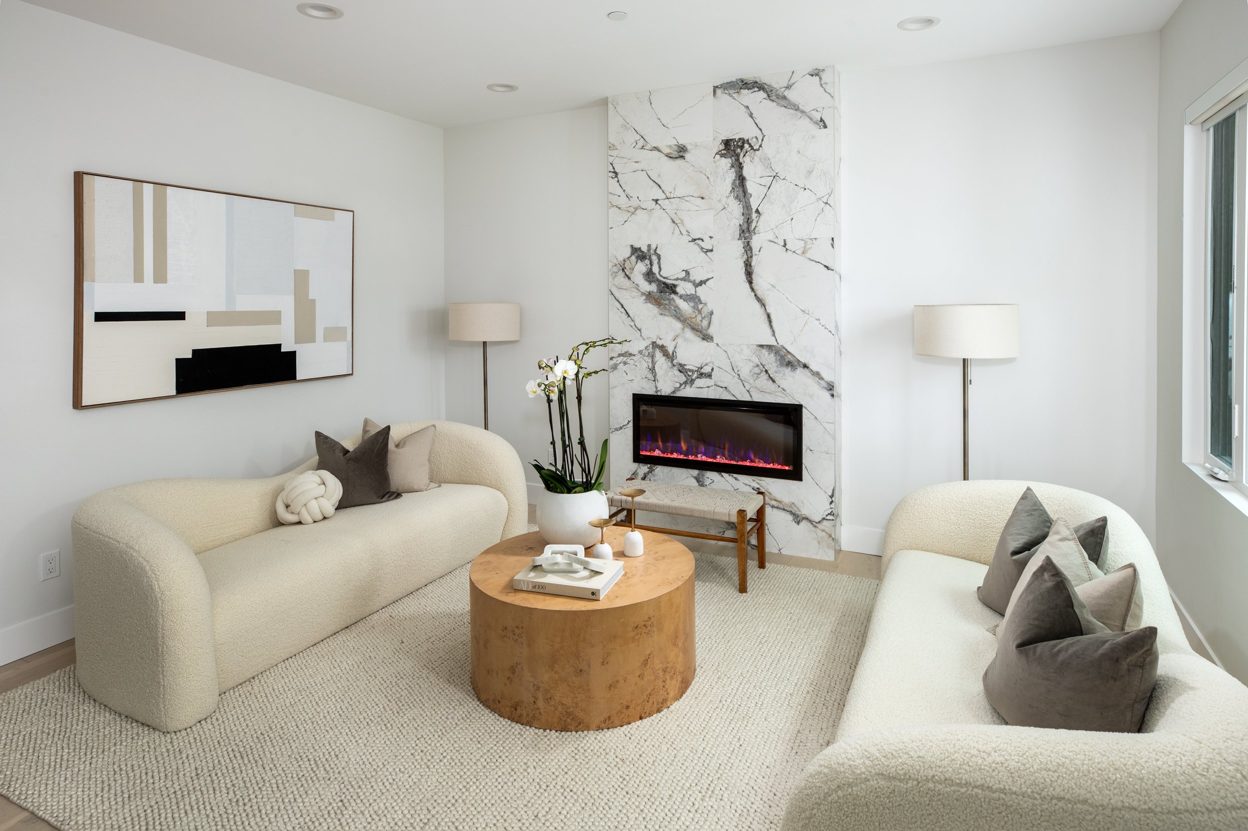 Living room with two cream-colored sofas, a round wooden coffee table, modern fireplace with marble surround, abstract wall art, and two tall floor lamps.