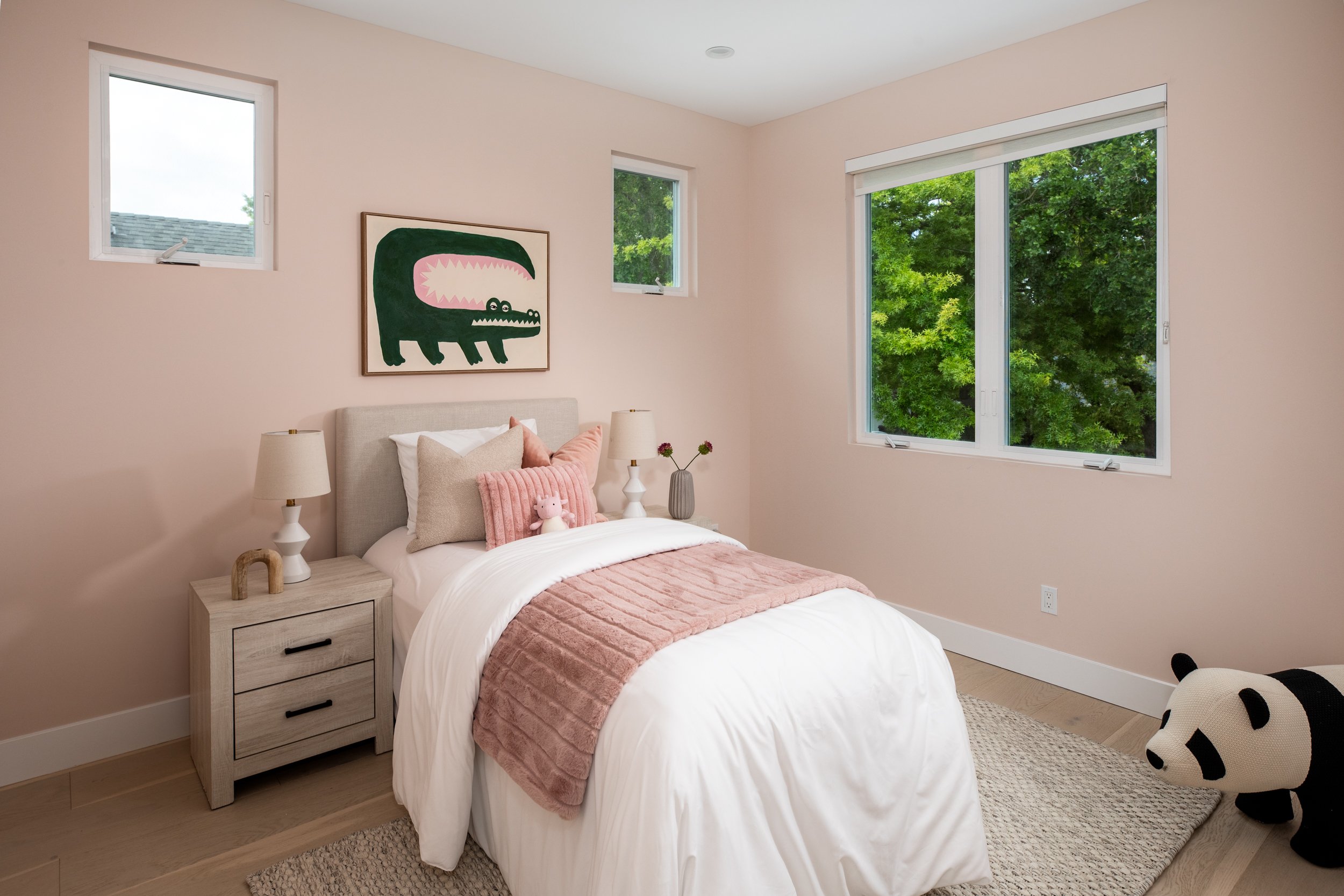 A bedroom with a bed, nightstand, and large window overlooking green trees. The bed has pink and beige pillows, a pink blanket, and a small plush toy. A framed crocodile picture hangs above the bed, and a panda stuffed toy is on the floor.