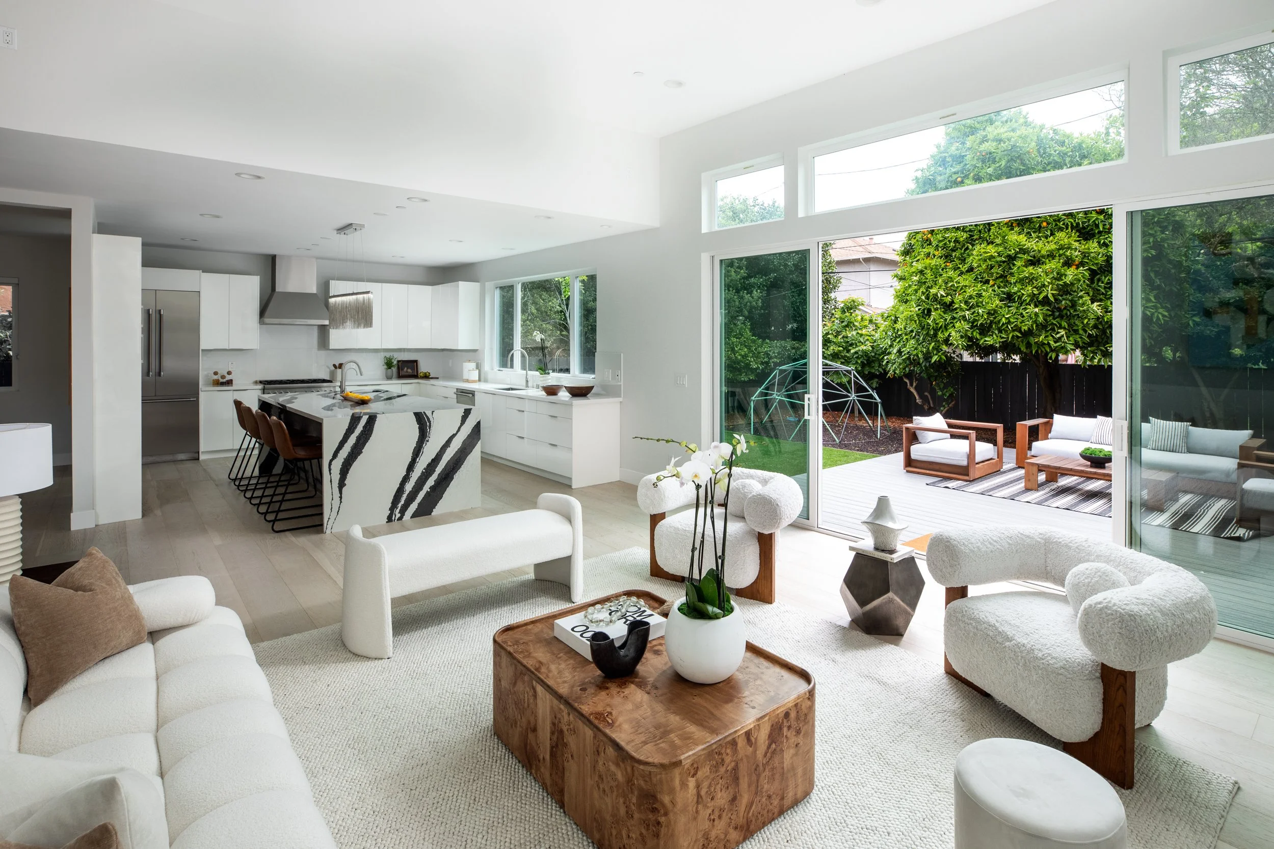 Bright modern living room with white sofas, wooden coffee table, and decorative vases. Open kitchen with white cabinets and a marble island. Large sliding glass door opens to outdoor patio with seating and greenery.