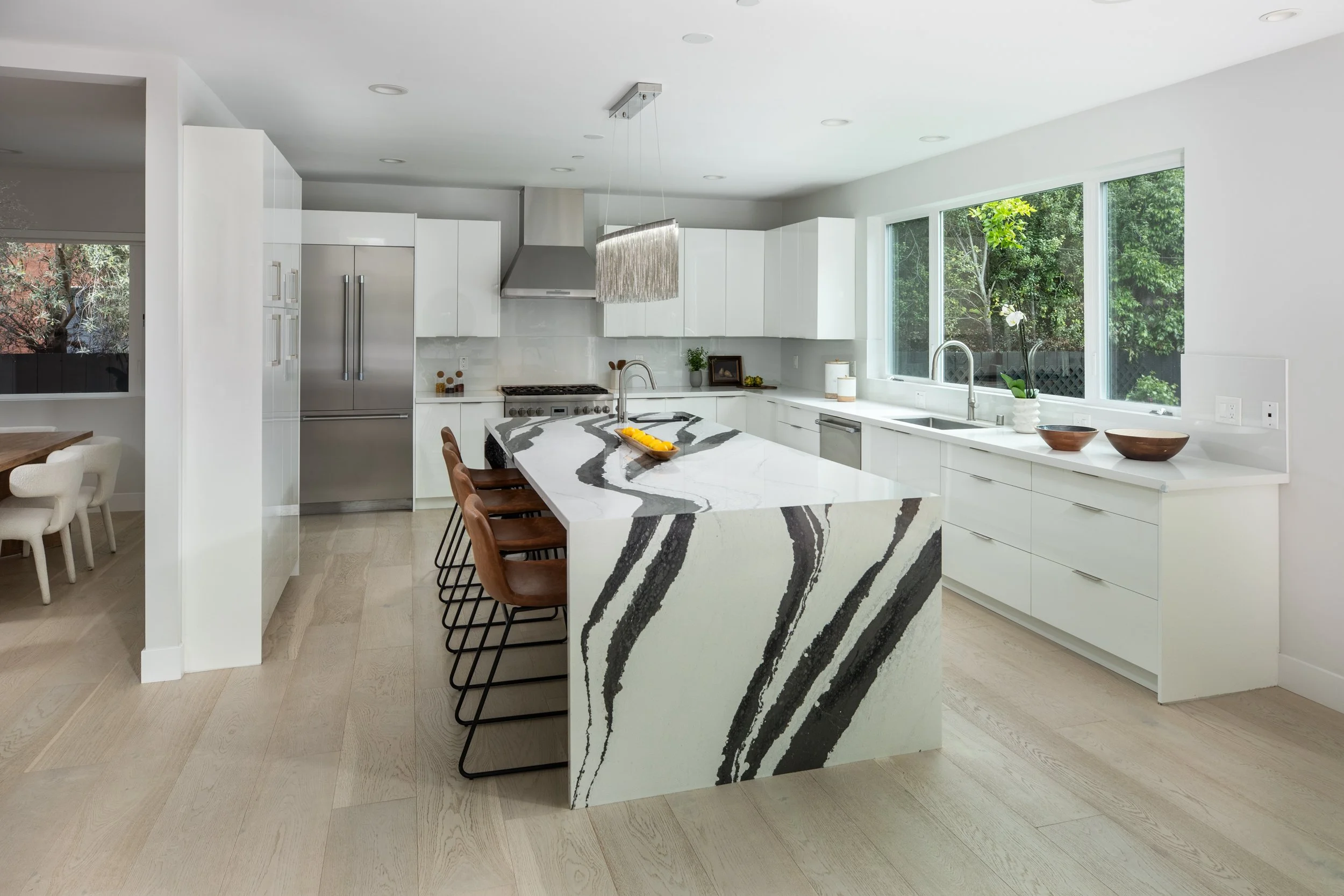 Modern kitchen with white cabinets, a marble island with black streaks, and brown chairs. Large window with greenery outside.