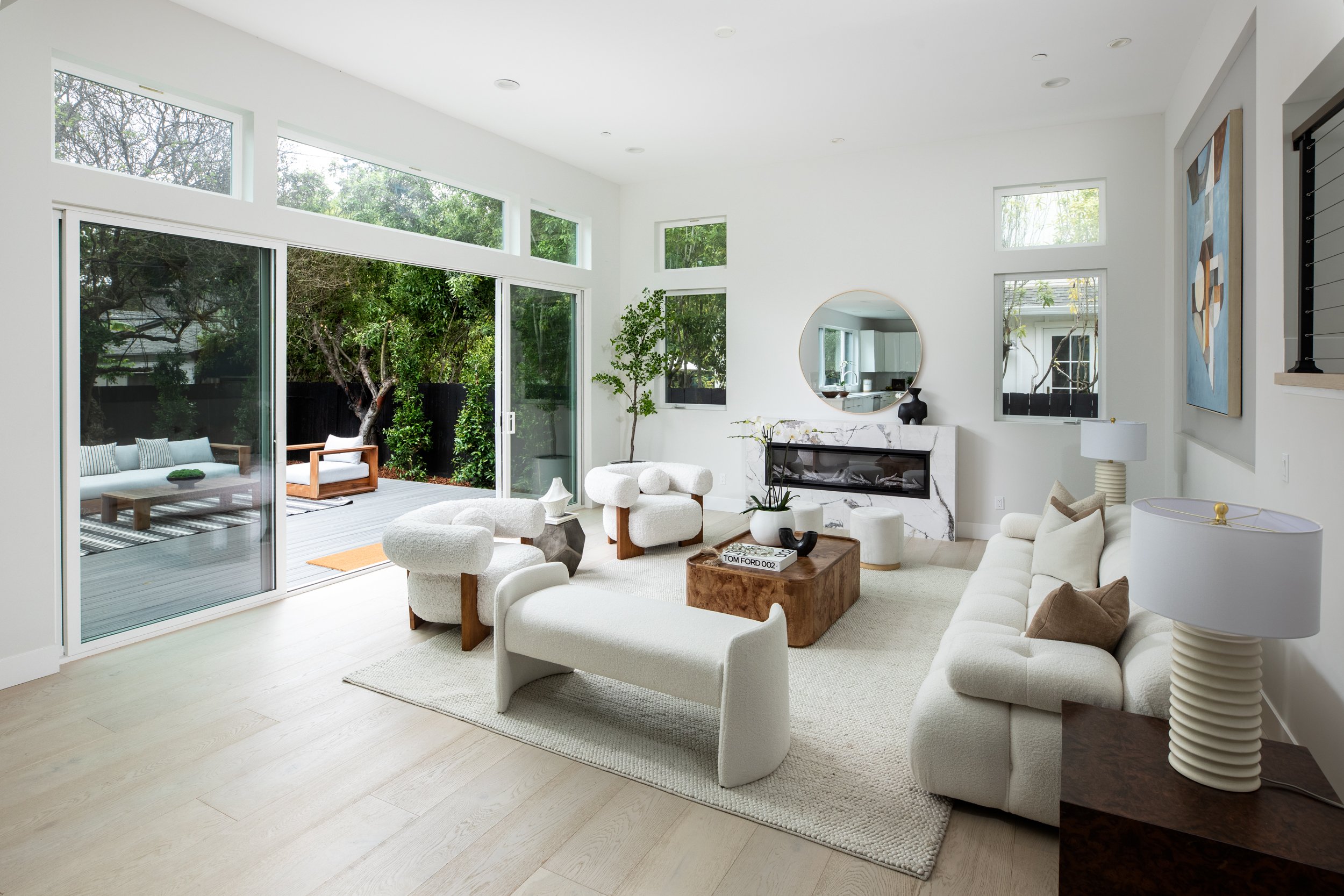 Modern living room with a large glass sliding door opening to an outdoor deck, minimalist white and beige sofas, wooden and marble accents, abstract art, and greenery outside
