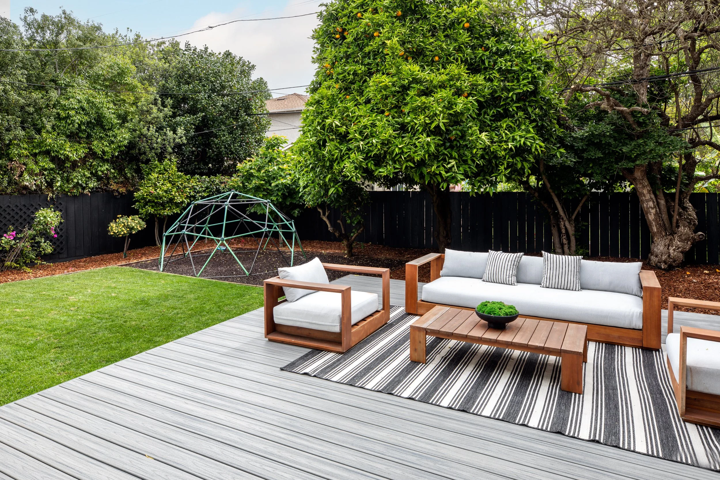 Outdoor patio with gray wooden deck, beige cushioned outdoor furniture, black and white striped rug, small black bowl with green plant, green trees, and a metal garden dome in backyard.