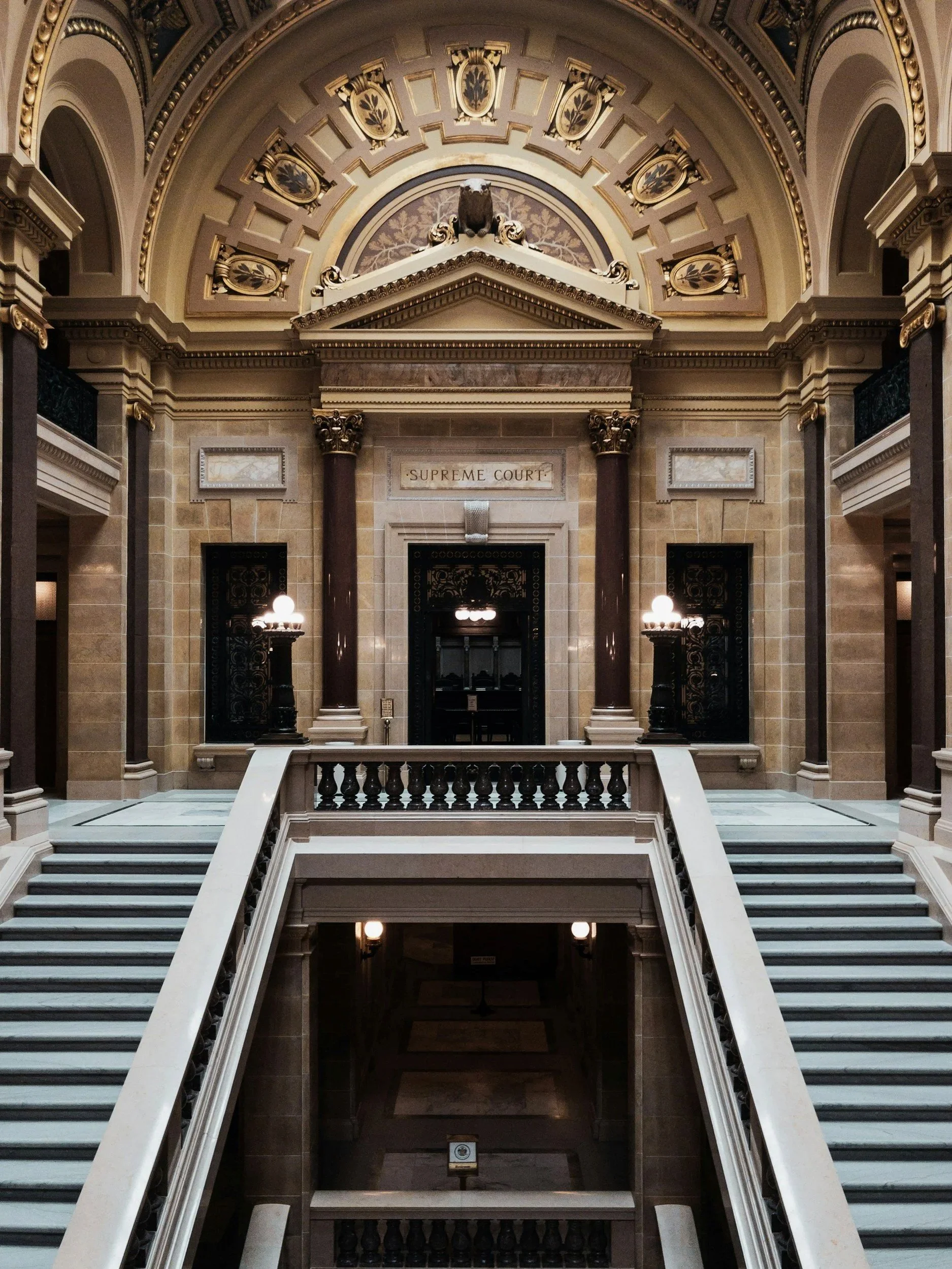 Interior of a government building with a staircase leading to a door labeled 'Supreme Court'. The architecture features marble columns, ornate moldings, and decorative ceiling details, including a bear sculpture above the doorway.
