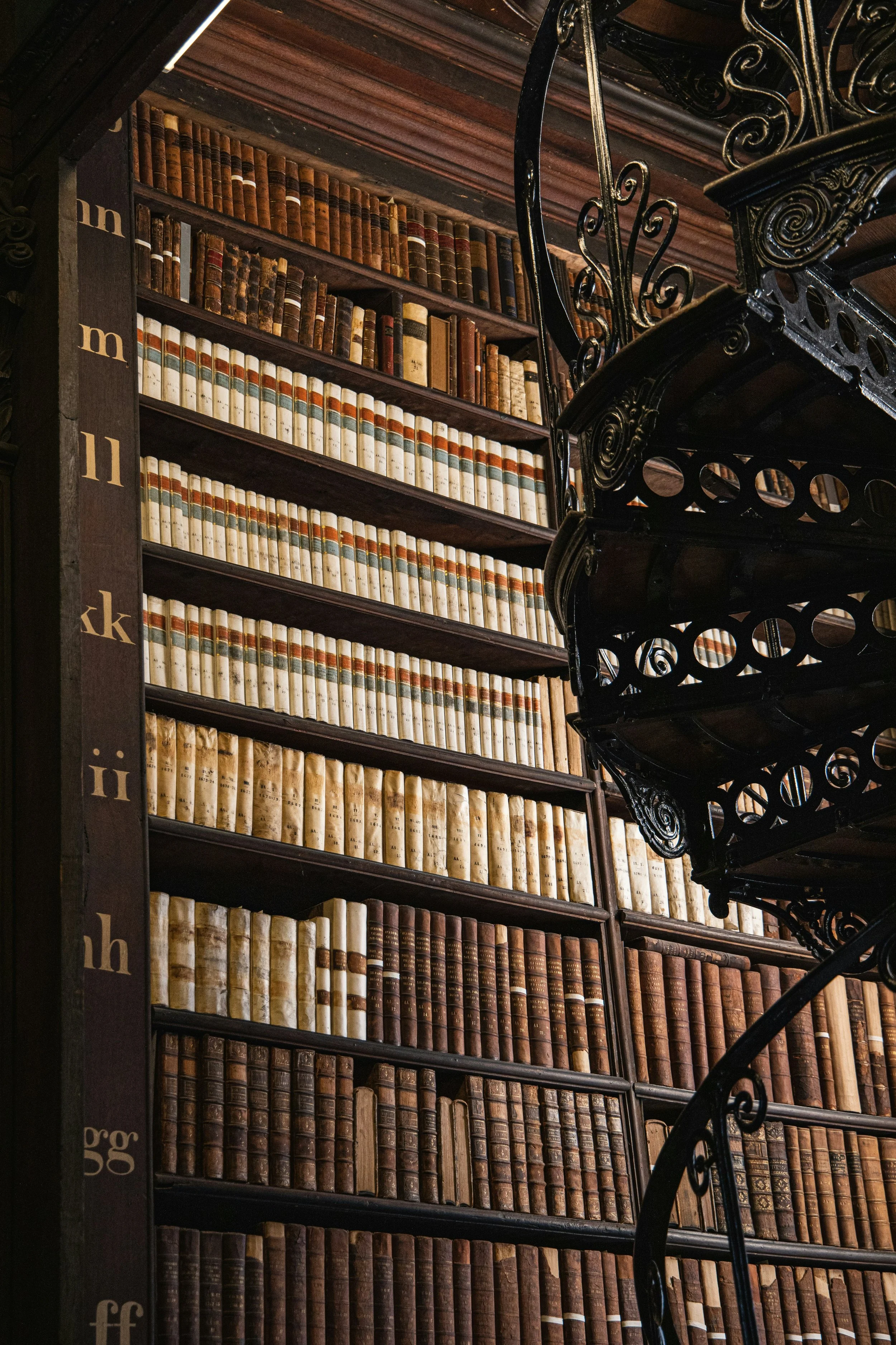 A tall wooden bookshelf filled with old, leather-bound books in a historical library. An ornate black iron spiral staircase is visible on the right side, partially obscuring some of the books.