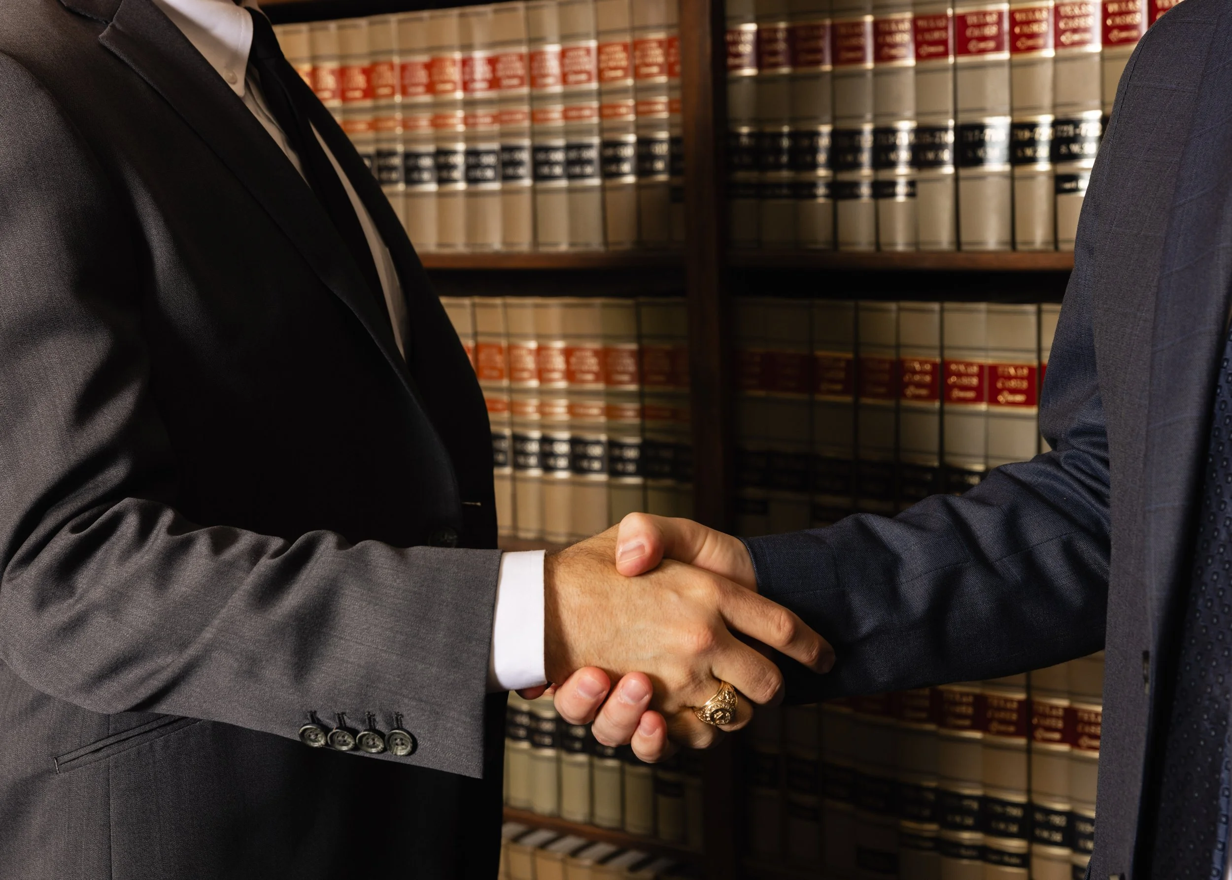 Two individuals in business suits shaking hands in a library or legal office, with shelves of law books in the background.