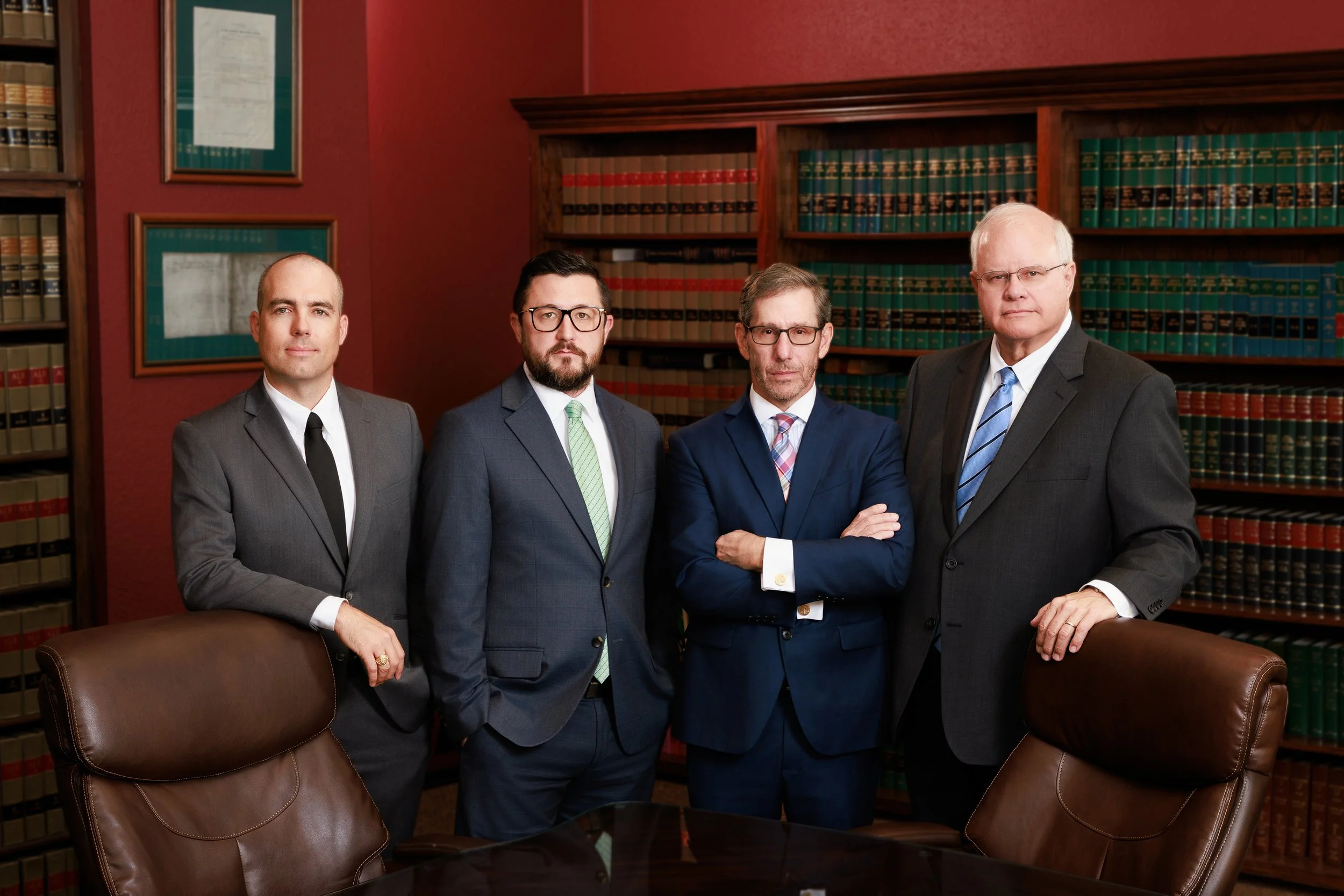 Four men in suits standing in a law office or library, with bookshelves filled with legal books in the background.