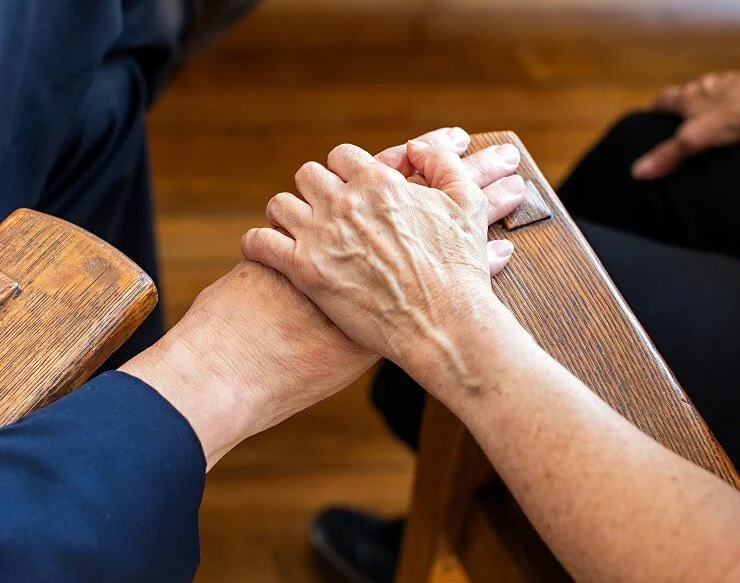 Two elderly people holding hands on a wooden table, one wearing a dark long sleeve shirt, in a warm indoor setting.