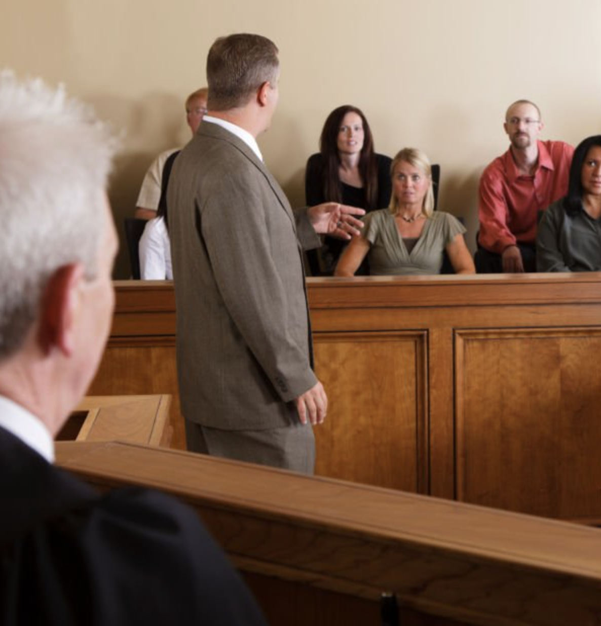 A man in a gray suit gives testimony in a courtroom, standing near the judge's bench while several people seated behind a wooden barrier listen attentively.