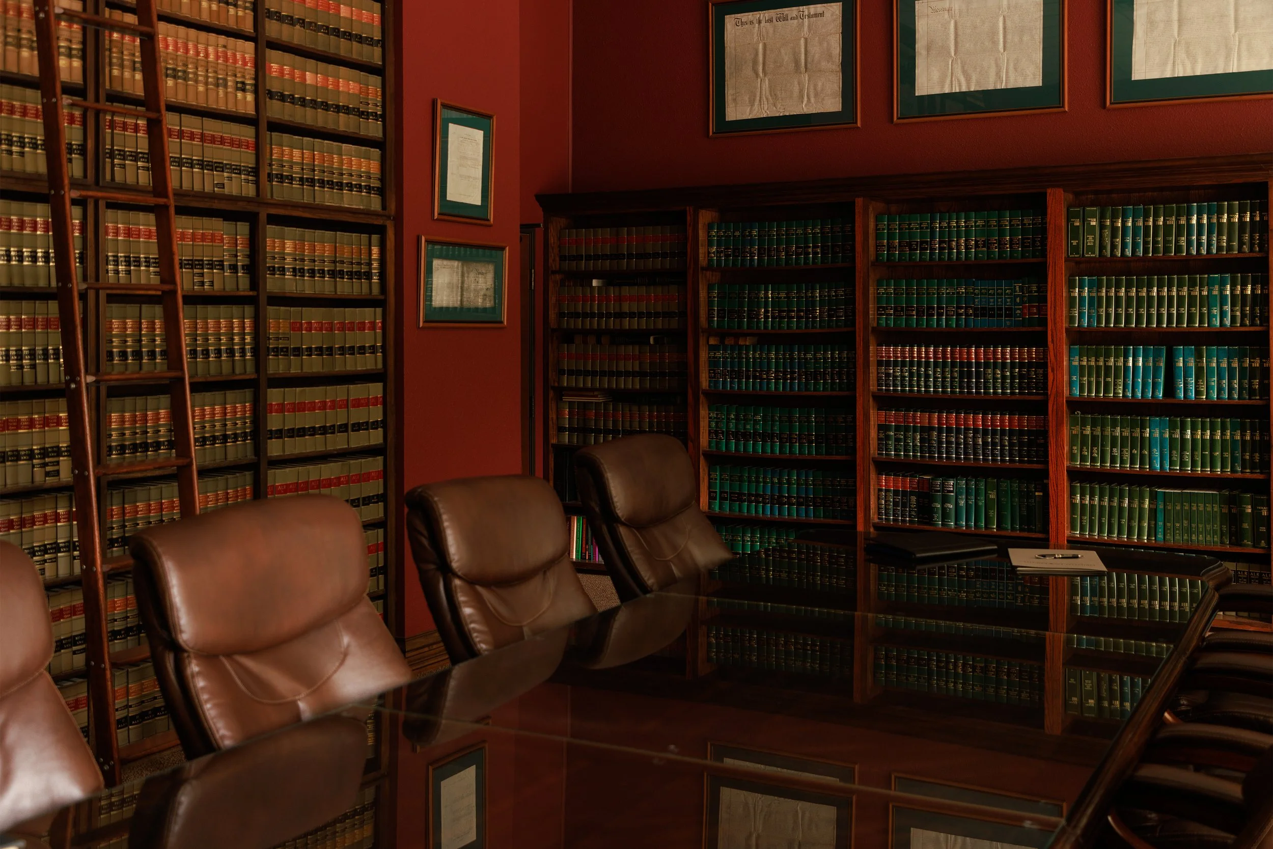 A conference room with leather chairs, wooden bookshelves filled with legal books, framed documents on the walls, and a reflective glass table.
