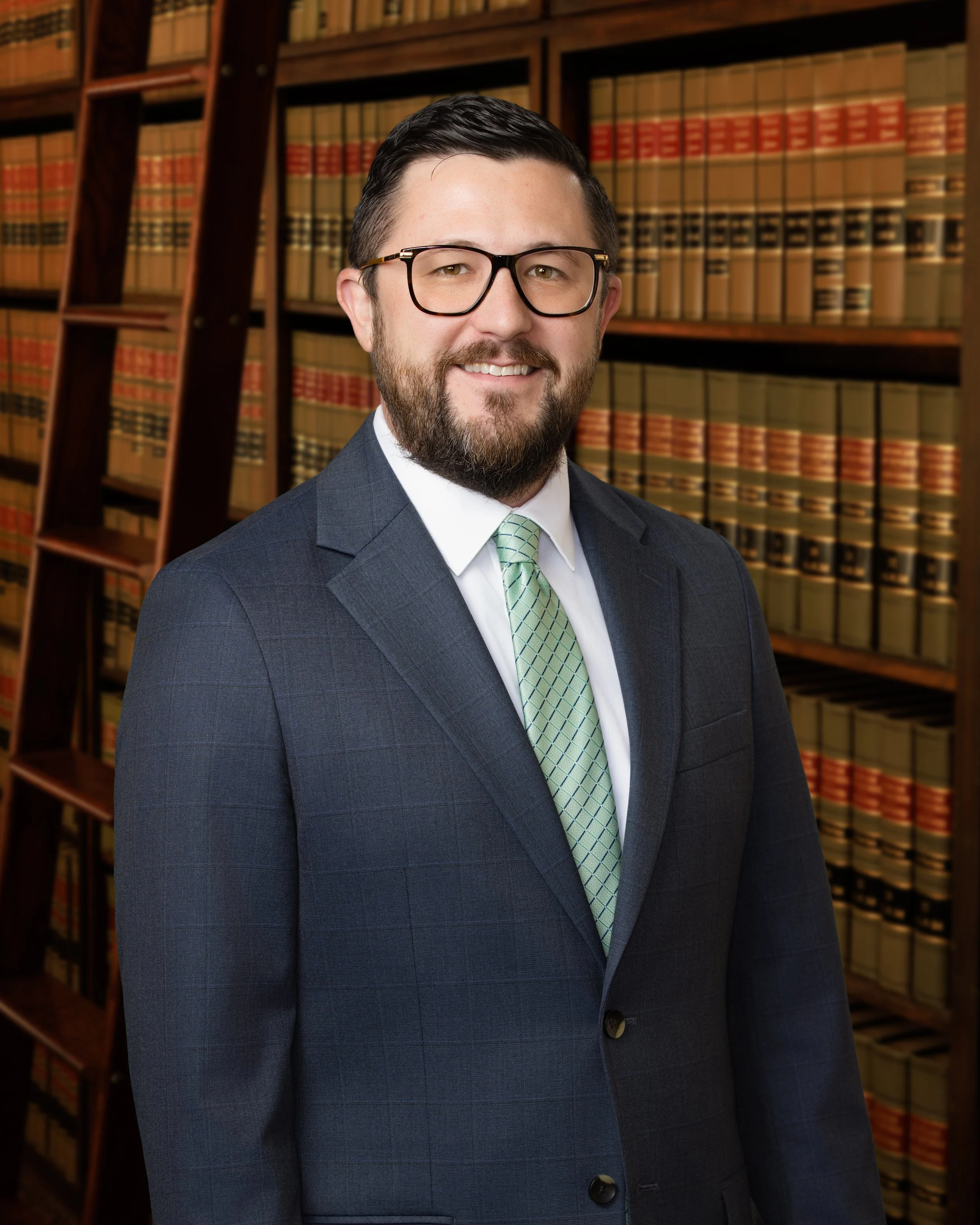 A man in a suit and tie wearing glasses, standing in front of bookshelves filled with law books in a library or office setting.