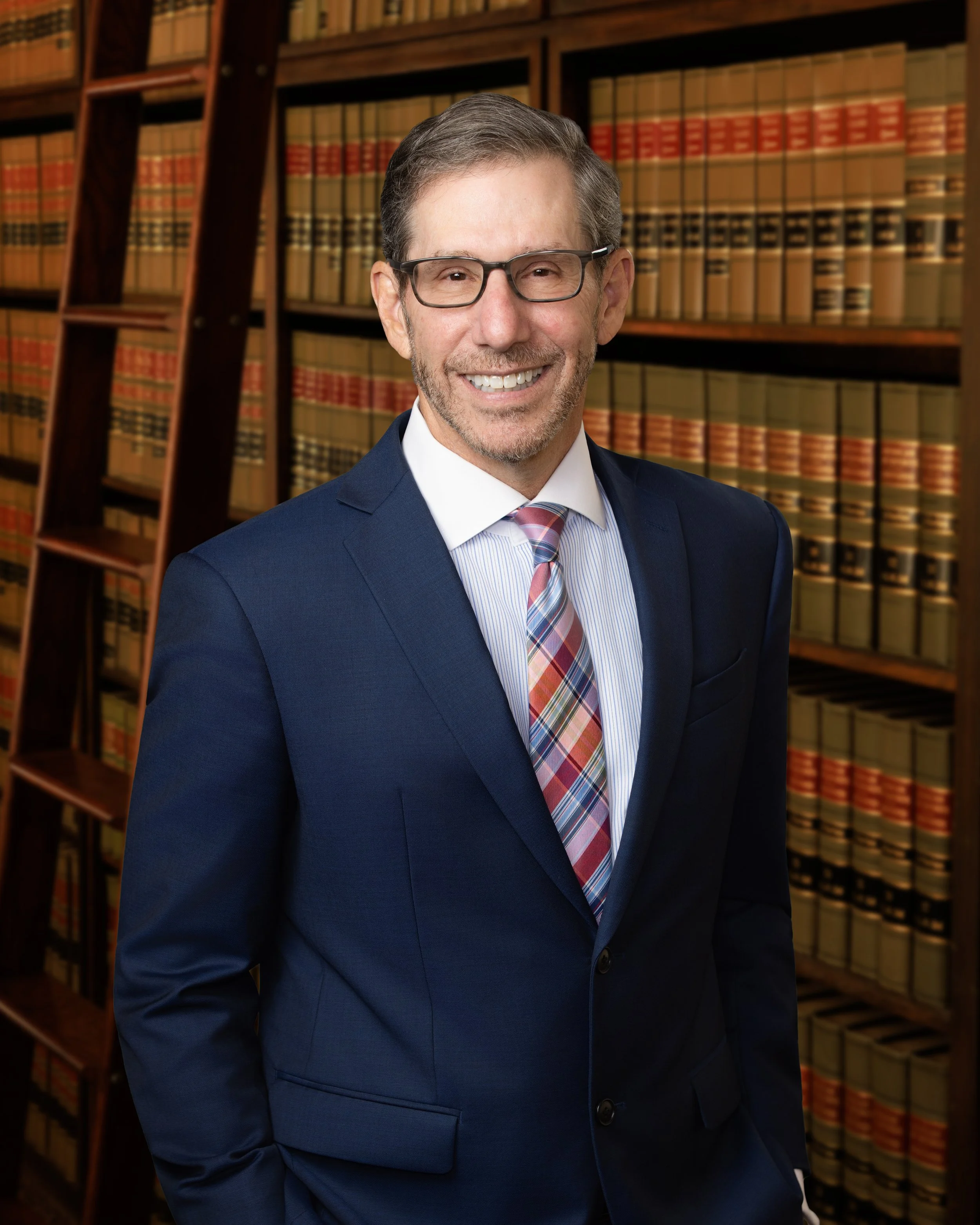 A professional man in a navy suit, light blue shirt, and pink plaid tie is smiling while standing in front of bookshelves filled with legal books in a library or office.