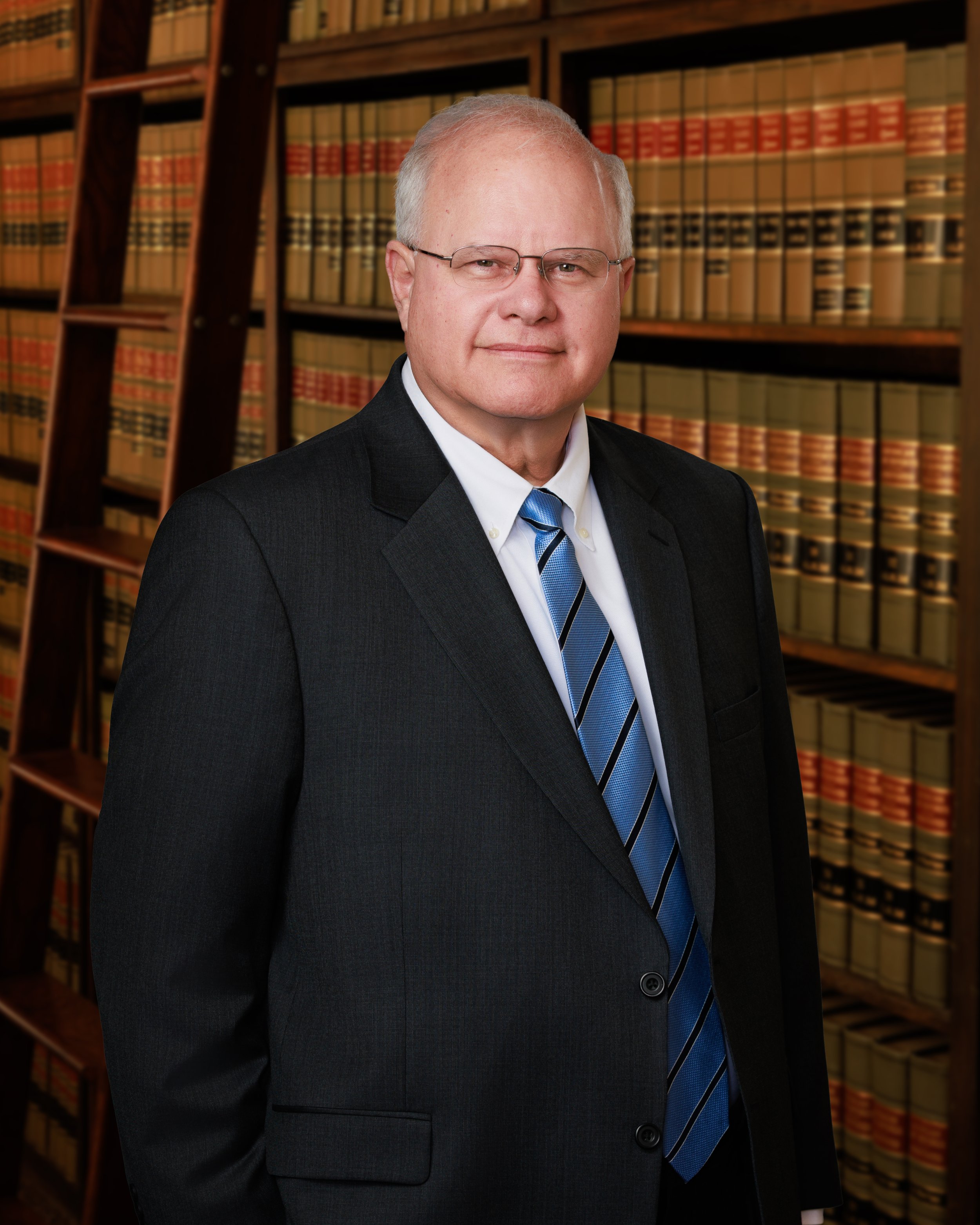 A man with glasses and light-colored hair wearing a dark suit, white shirt, and blue striped tie standing in front of shelves filled with legal books in a law library.