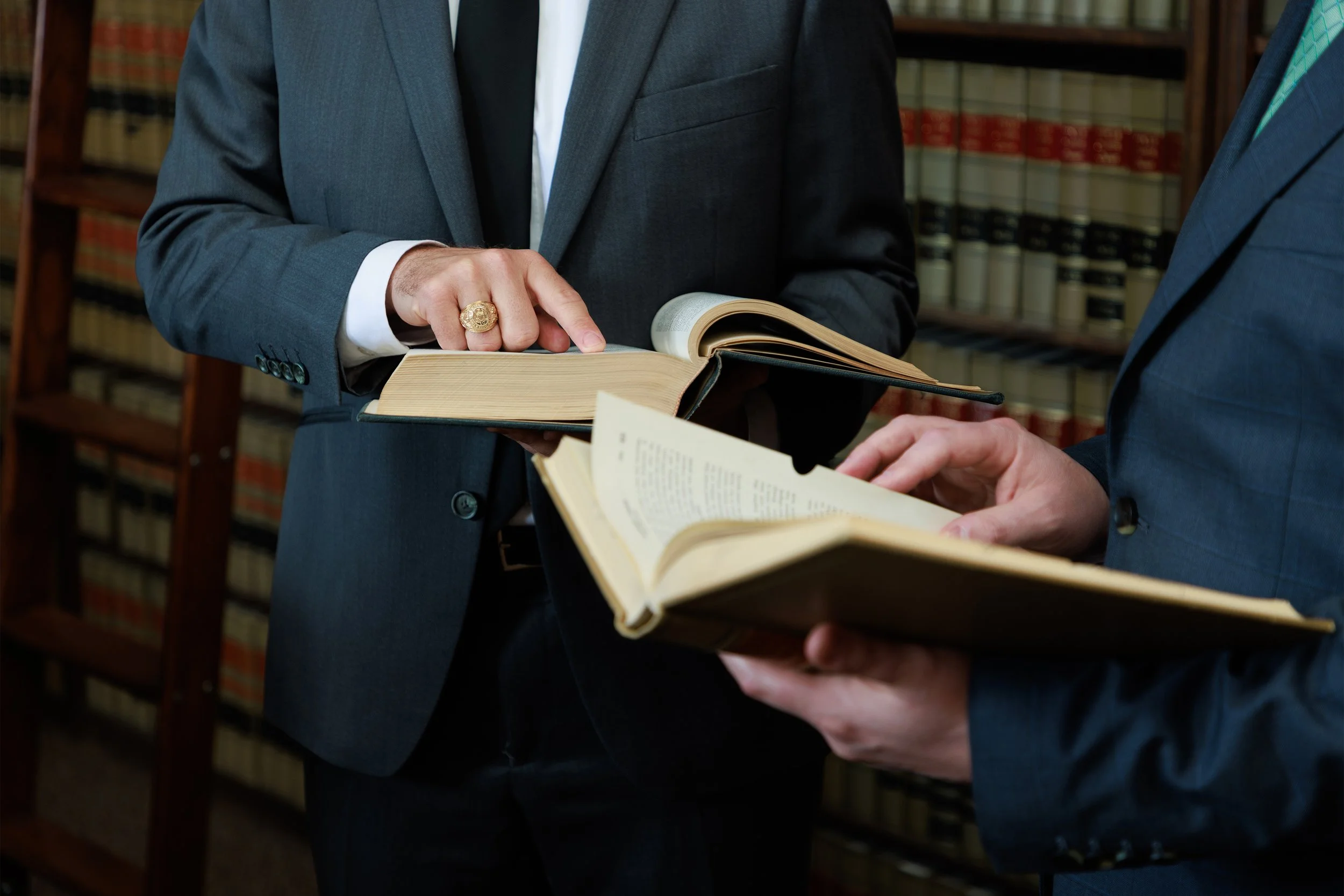 Two people in formal suits are examining a large open book in a library with shelves of legal books in the background.