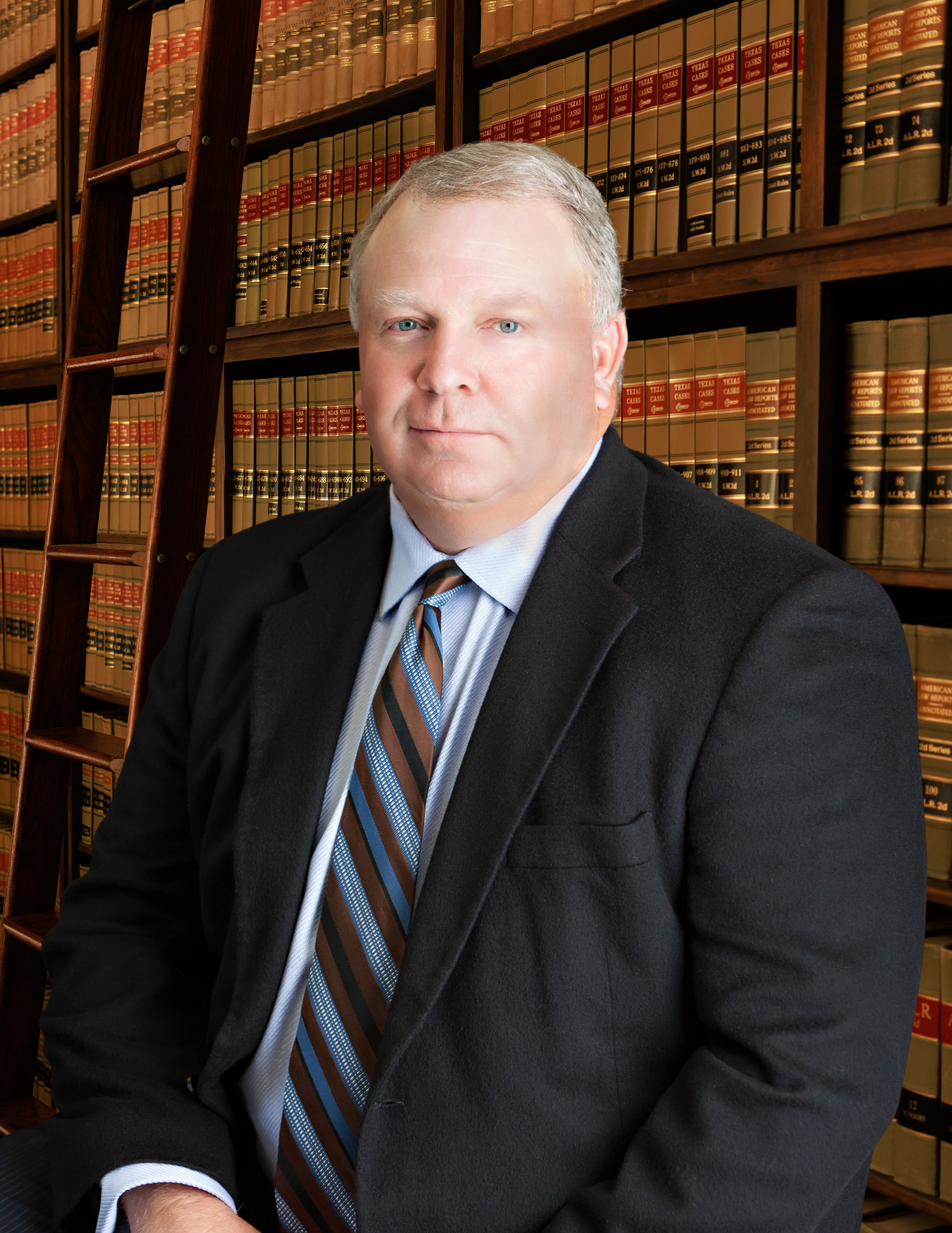 Close-up of a man in a suit with a striped tie, standing in front of a large wooden bookshelf filled with law books.