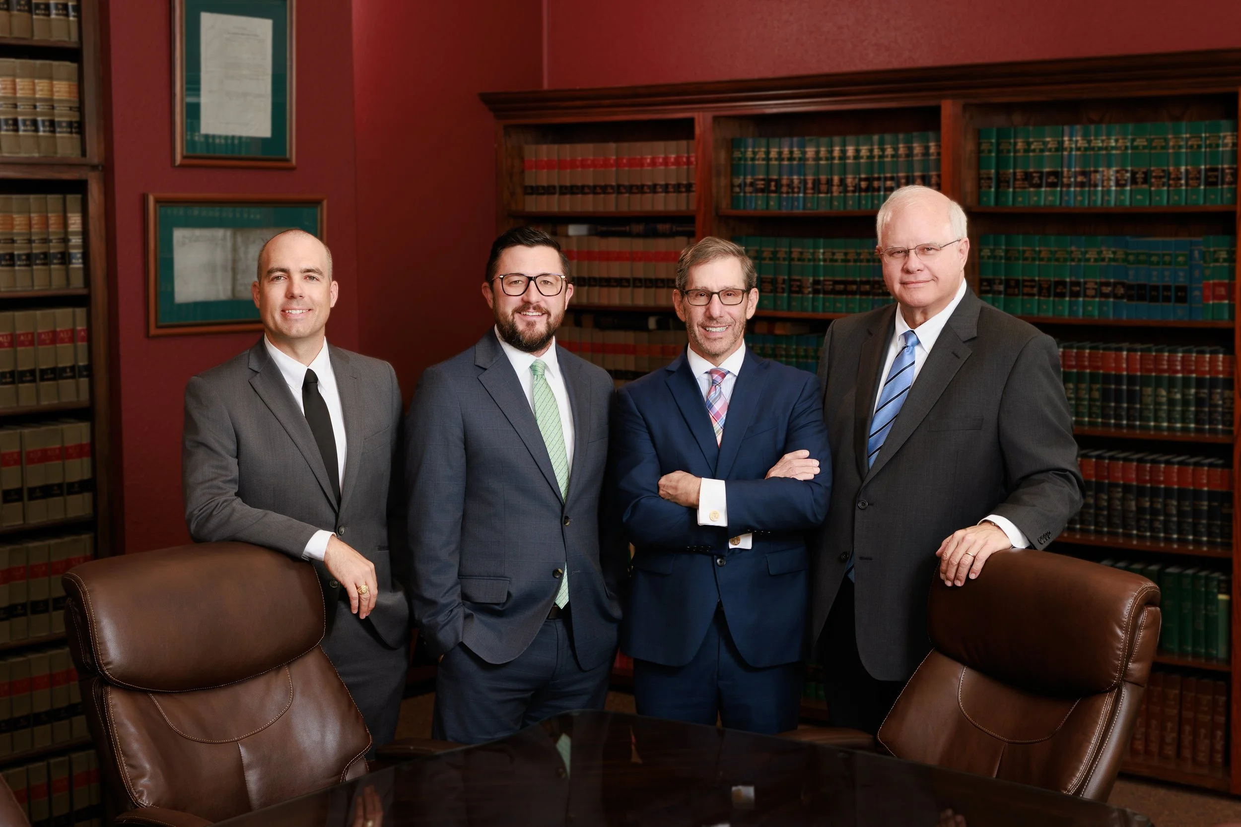 Four men in suits standing in a law office or library, with bookshelves filled with legal books behind them and framed certificates or documents on the wall.