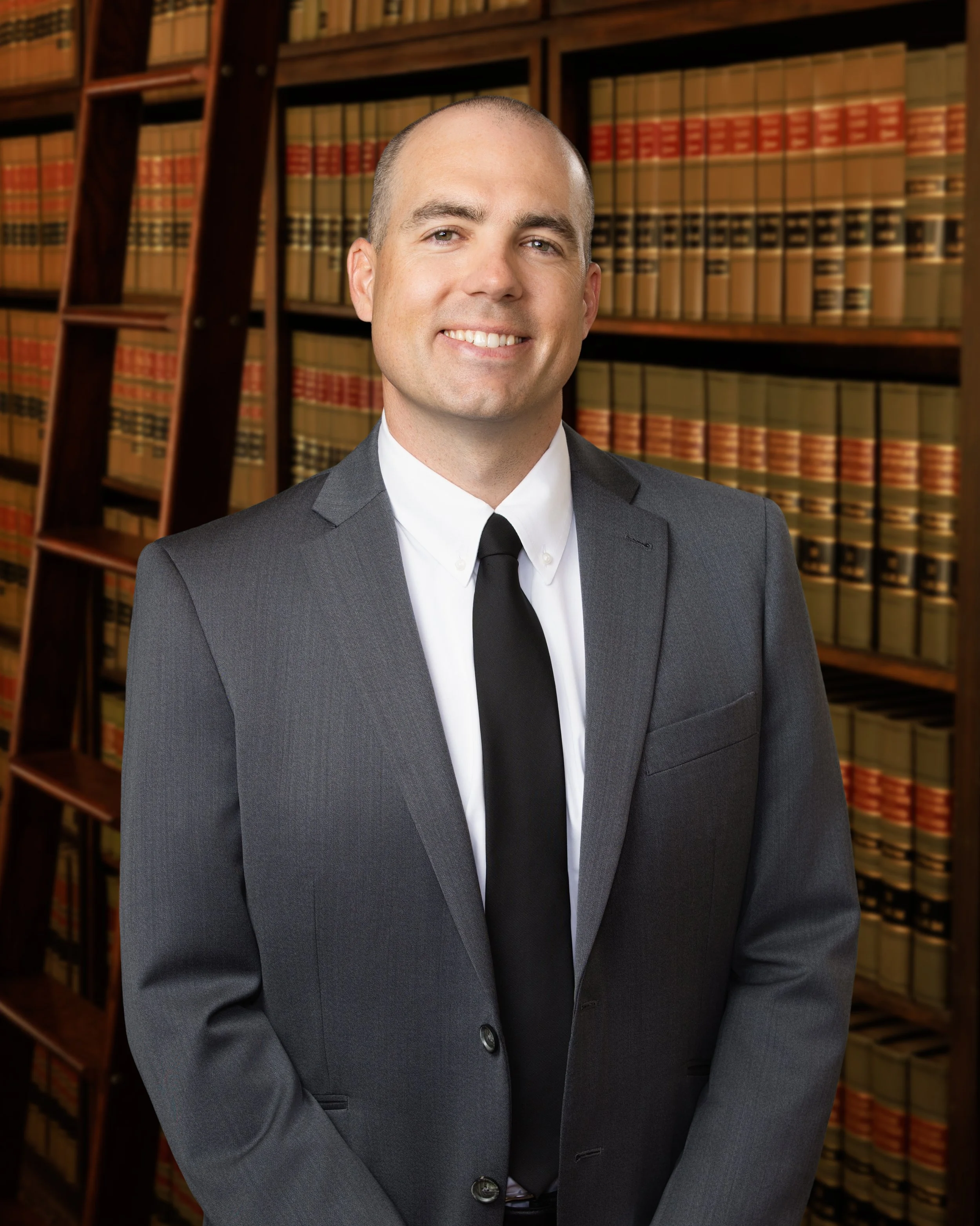 A man in a gray suit smiling in front of a bookshelf filled with legal books.