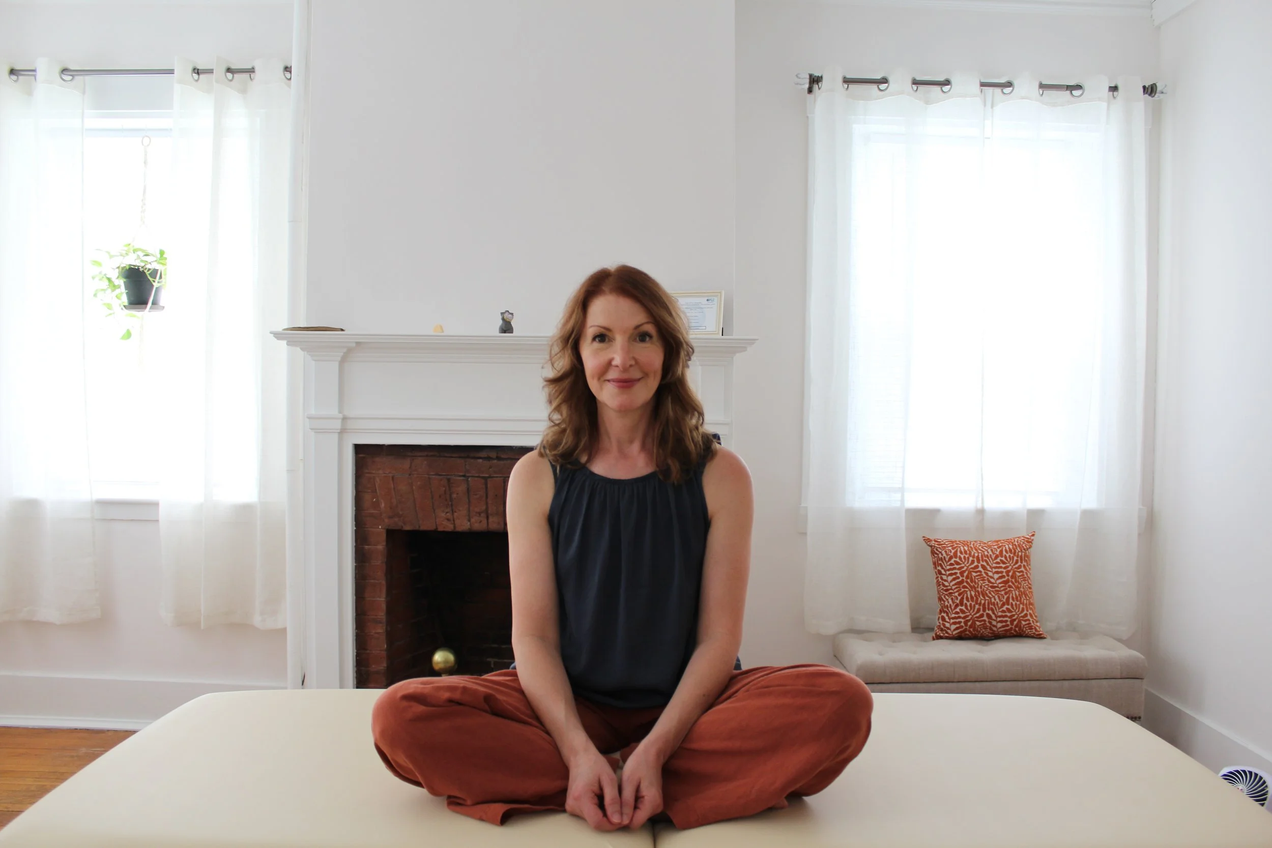 A woman with shoulder-length auburn hair smiling while sitting cross-legged on a white table in a bright living room with white walls, sheer curtains, a brick fireplace, and a red patterned pillow on a beige bench.