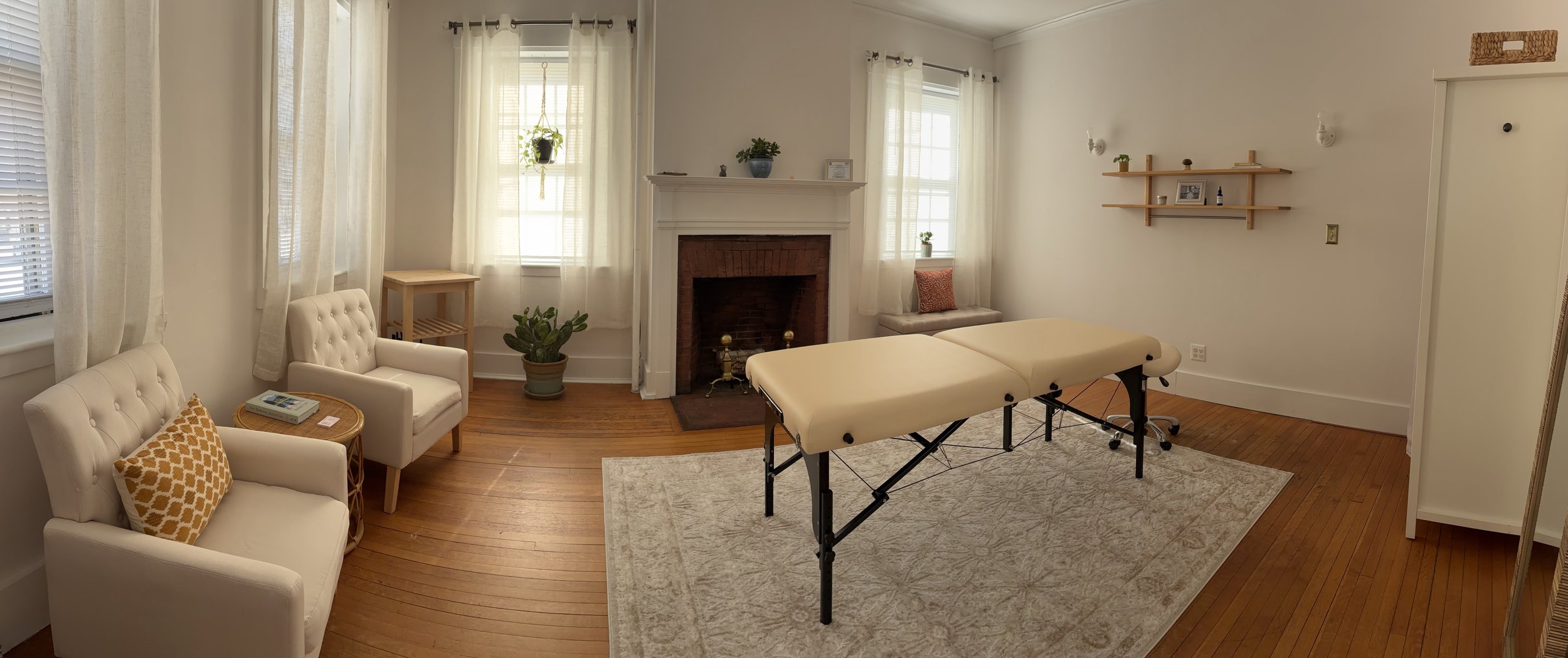 A cozy living room with white walls and hardwood floors featuring a massage table in the center, a fireplace with a white mantel, two windows with sheer curtains, two white armchairs with a small side table, a green potted plant, wall-mounted shelves with decorative items, and a mirror.