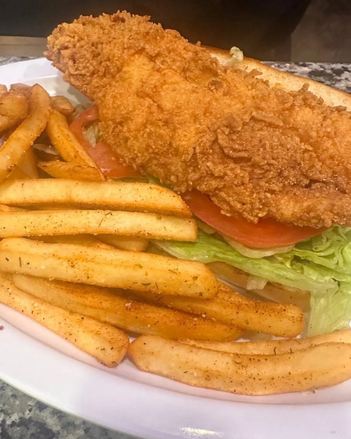 Fried chicken sandwich with lettuce, tomato, pickles, and French fries on a white plate.