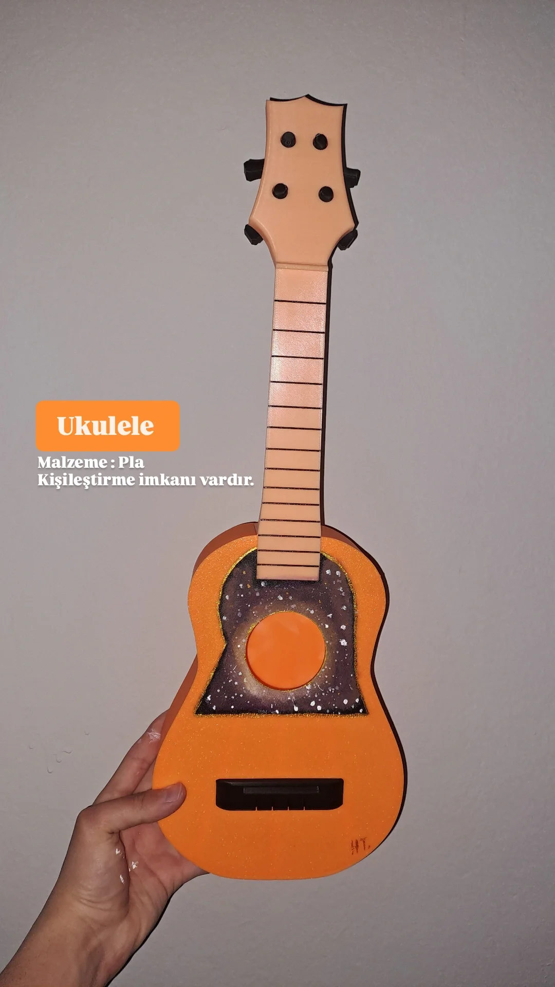 Hand holding a colorful, artistic wooden ukulele with a space-themed design on the body and a plain upright neck, against a plain background.