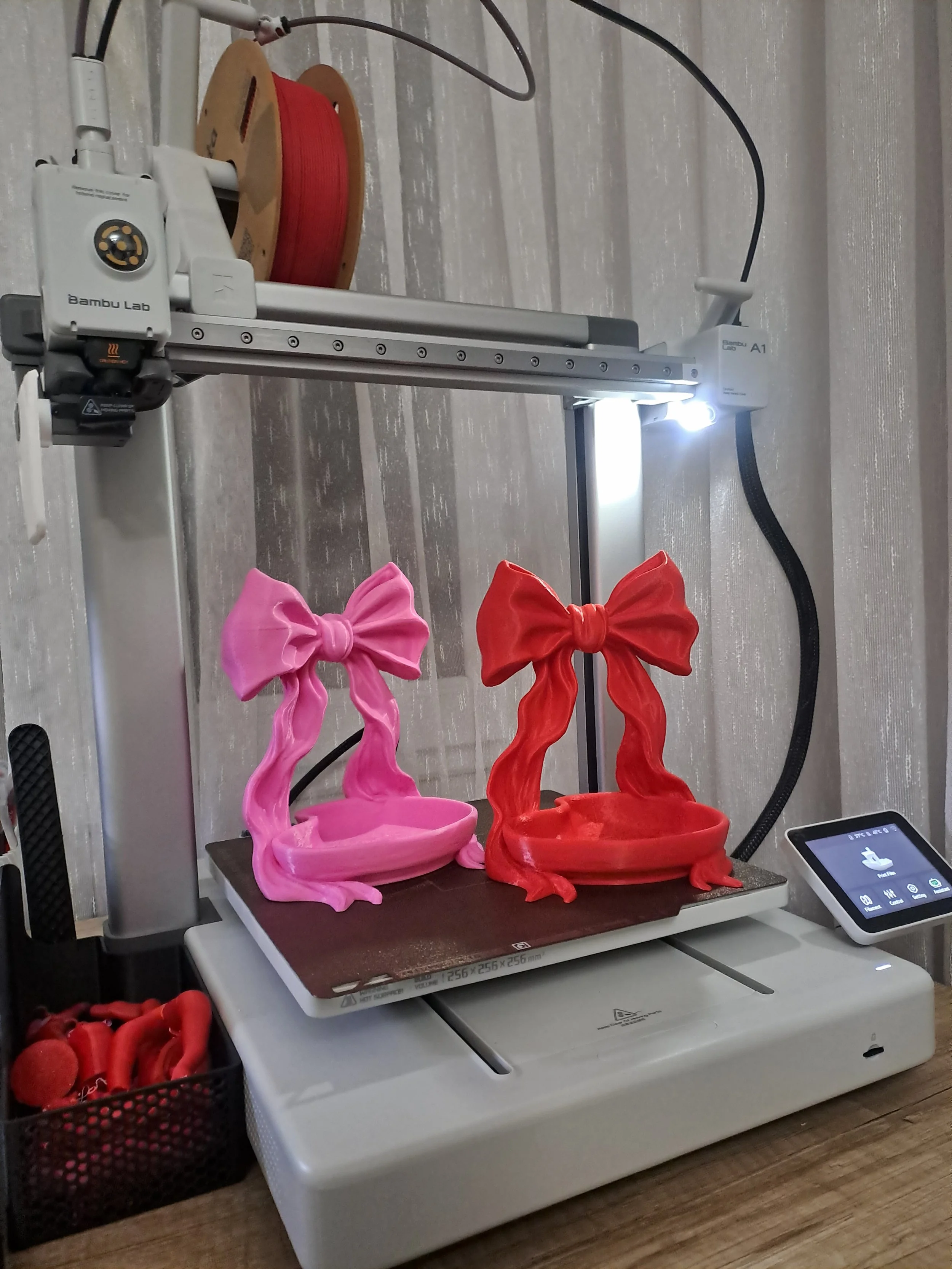 A 3D printer with two colorful bow-shaped objects, one pink and one red, on its build platform. The printer's extruder is active, and a control screen is visible to the right. There are also spools of filament and tools nearby.