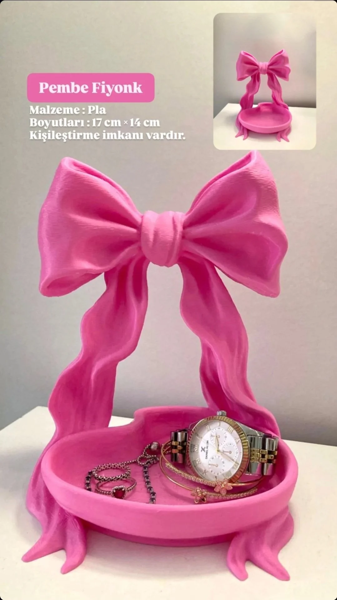 Pink decorative jewelry stand shaped like a bow with bracelets and necklaces on it, displaying a watch. The stand is made of pink resin and is 17 cm by 14 cm, with a bow design at the top. The background includes a small inset image showing the same jewelry holder from a different angle.