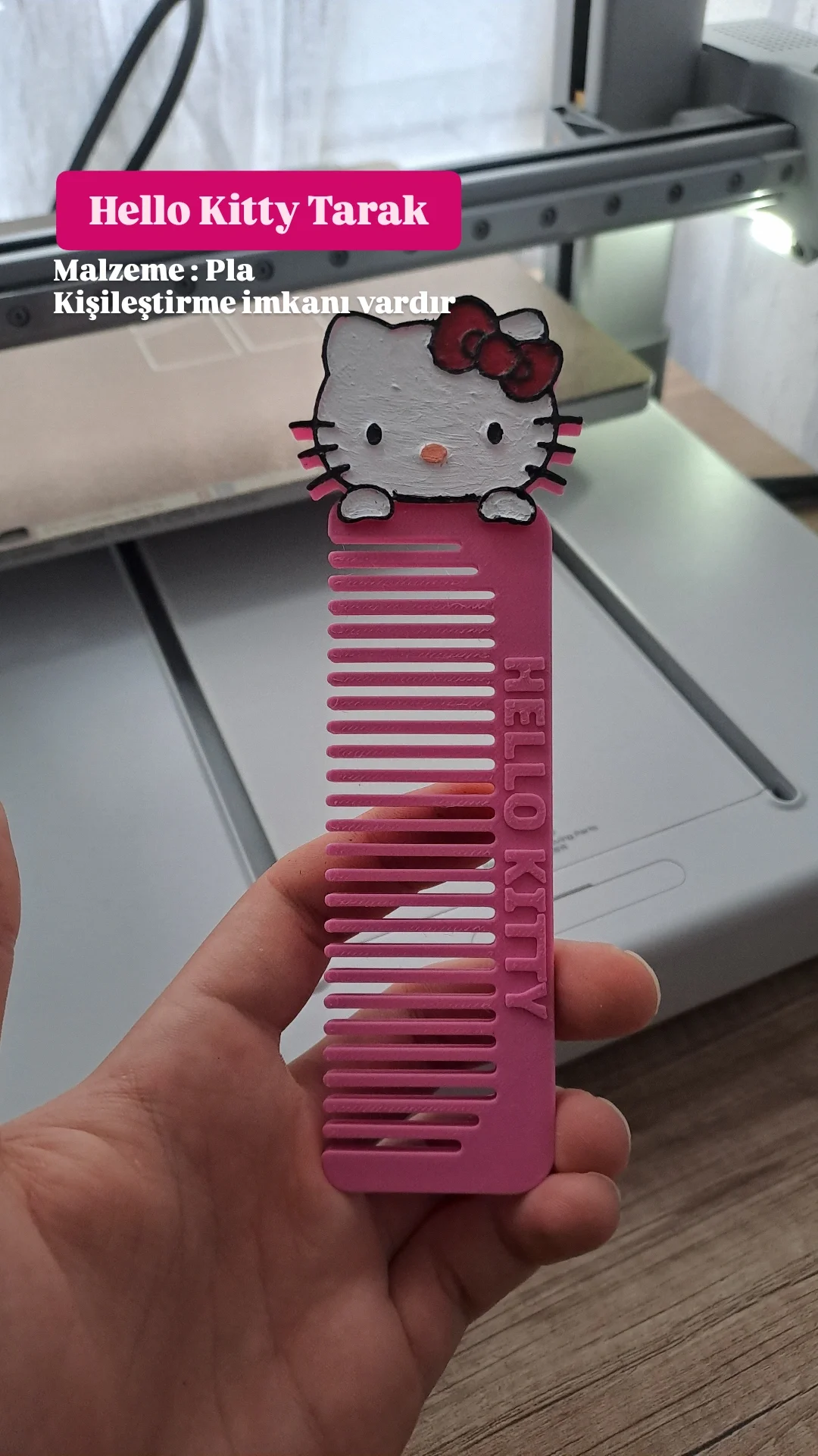 A pink Hello Kitty themed hair comb with a figure of Hello Kitty's face on top, held in front of a gray printer.