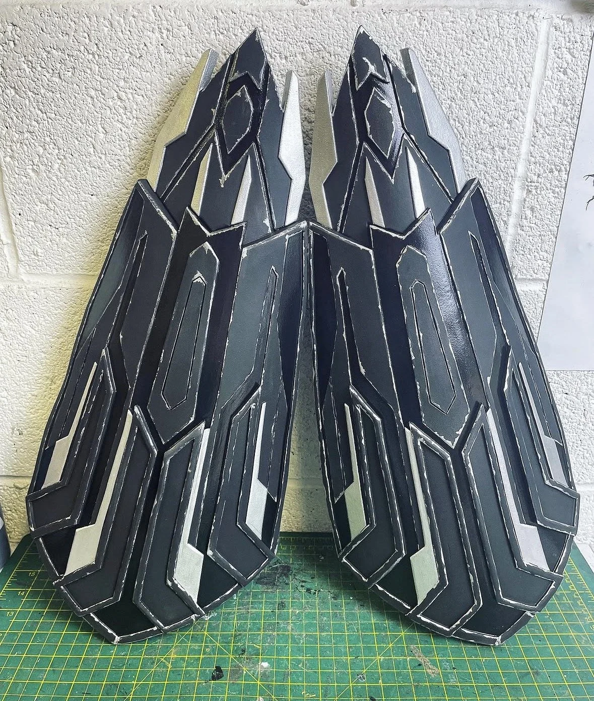 A large, black, and silver sculptural piece with angular geometric patterns leaning against a textured white wall, placed on a green cutting mat.
