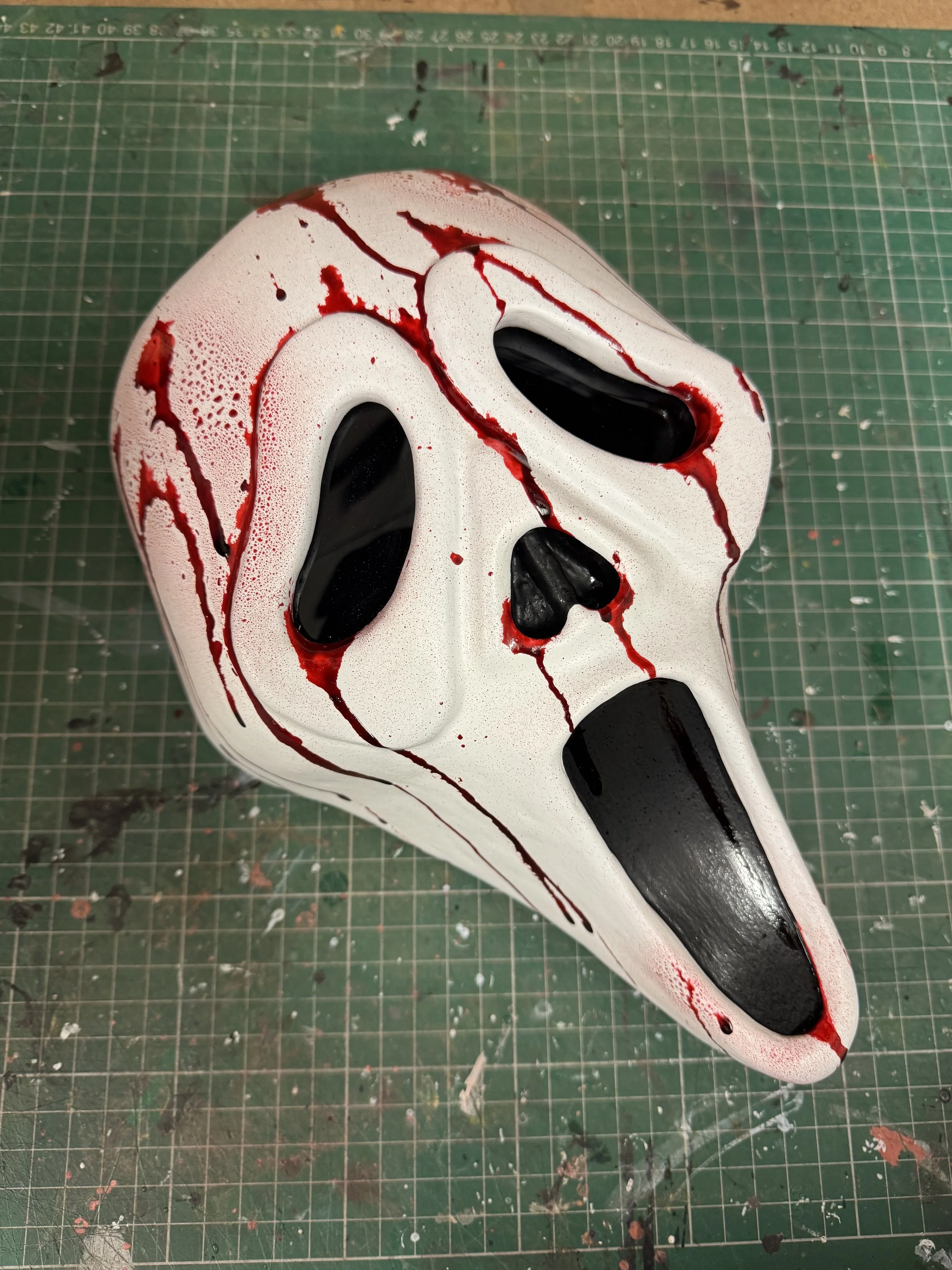 A sculpted mask resembling a ghost face with a white base, black eye holes, and red streaks for blood, placed on a green cutting mat with grid lines and blood splatters.