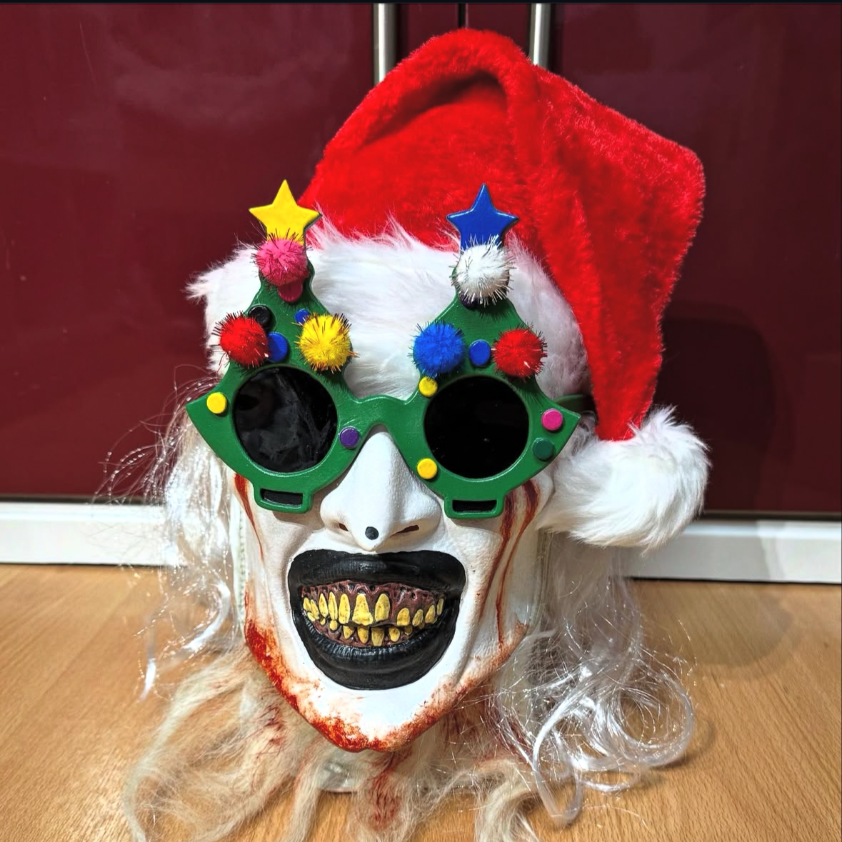 A creepy clown mask wearing a red Santa hat, decorated with festive Christmas glasses, and having white curly hair and a painted face with an open mouth showing yellow teeth and black lips.