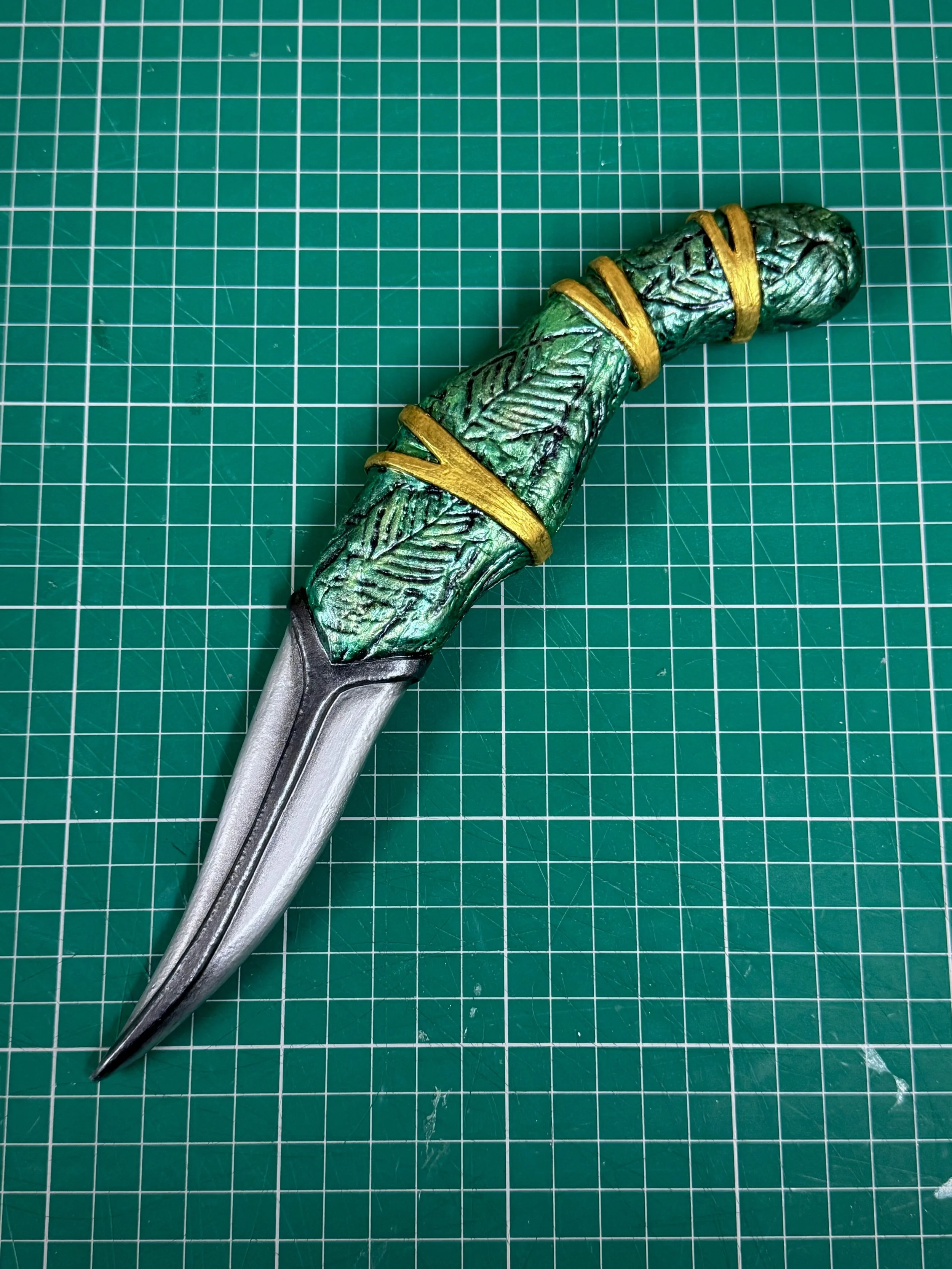 A foam prop dagger with a handle wrapped in green and gold decorative material, resting on a green cutting mat with white grid lines.