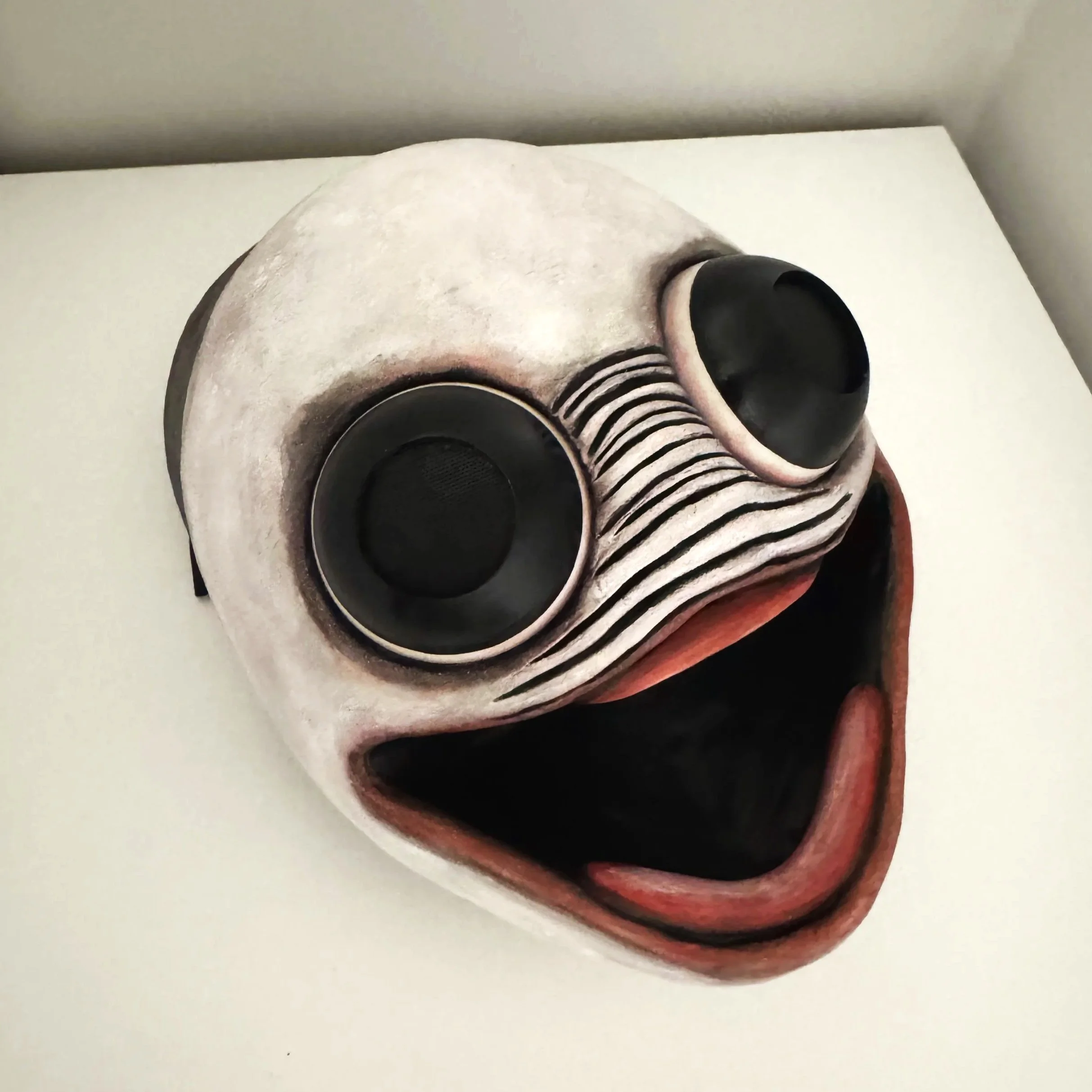 Close-up of a creepy mask with black eye sockets, a nose, and a painted mouth, placed on a white surface with a gray wall in the background.