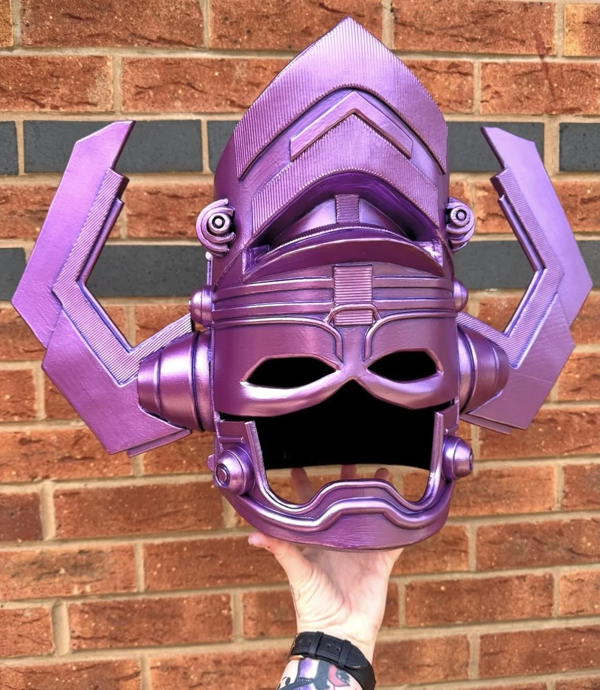 A purple metallic mask resembling Galactus from Fantastic 4 featuring an intricate design, held in front of a brick wall.