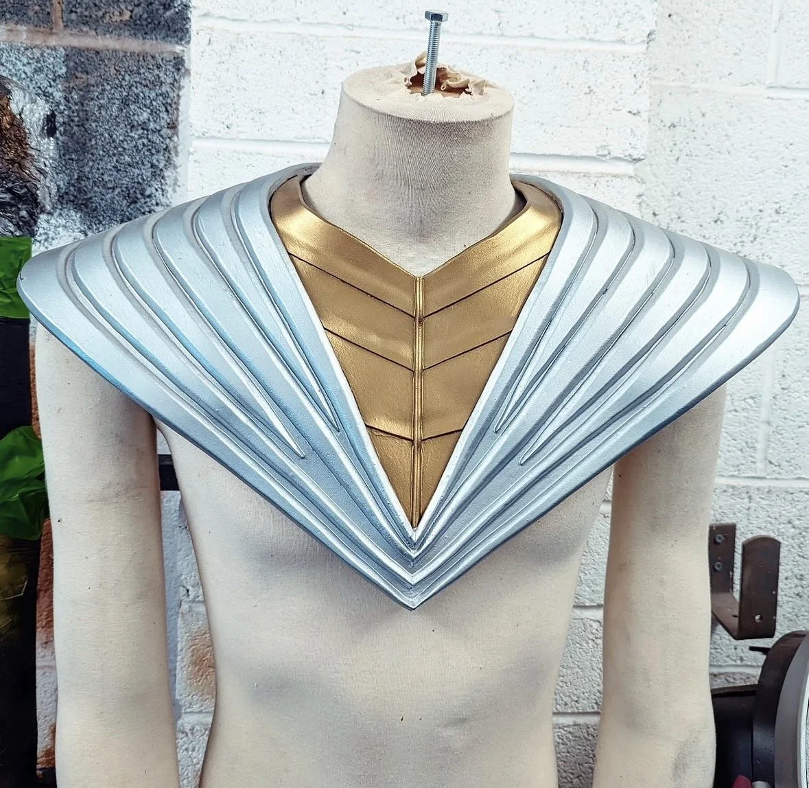 A mannequin wearing a futuristic costume with a gold chest piece and silver, fan-like shoulder armor against a white brick wall.