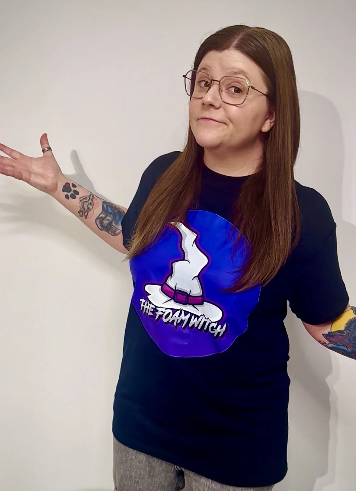 A woman with long brown hair, glasses and tattoos on her arms, wearing a black t-shirt with a graphic of a witches hat and the text 'The Foam Witch,' standing against a plain white wall with her arms outstretched.