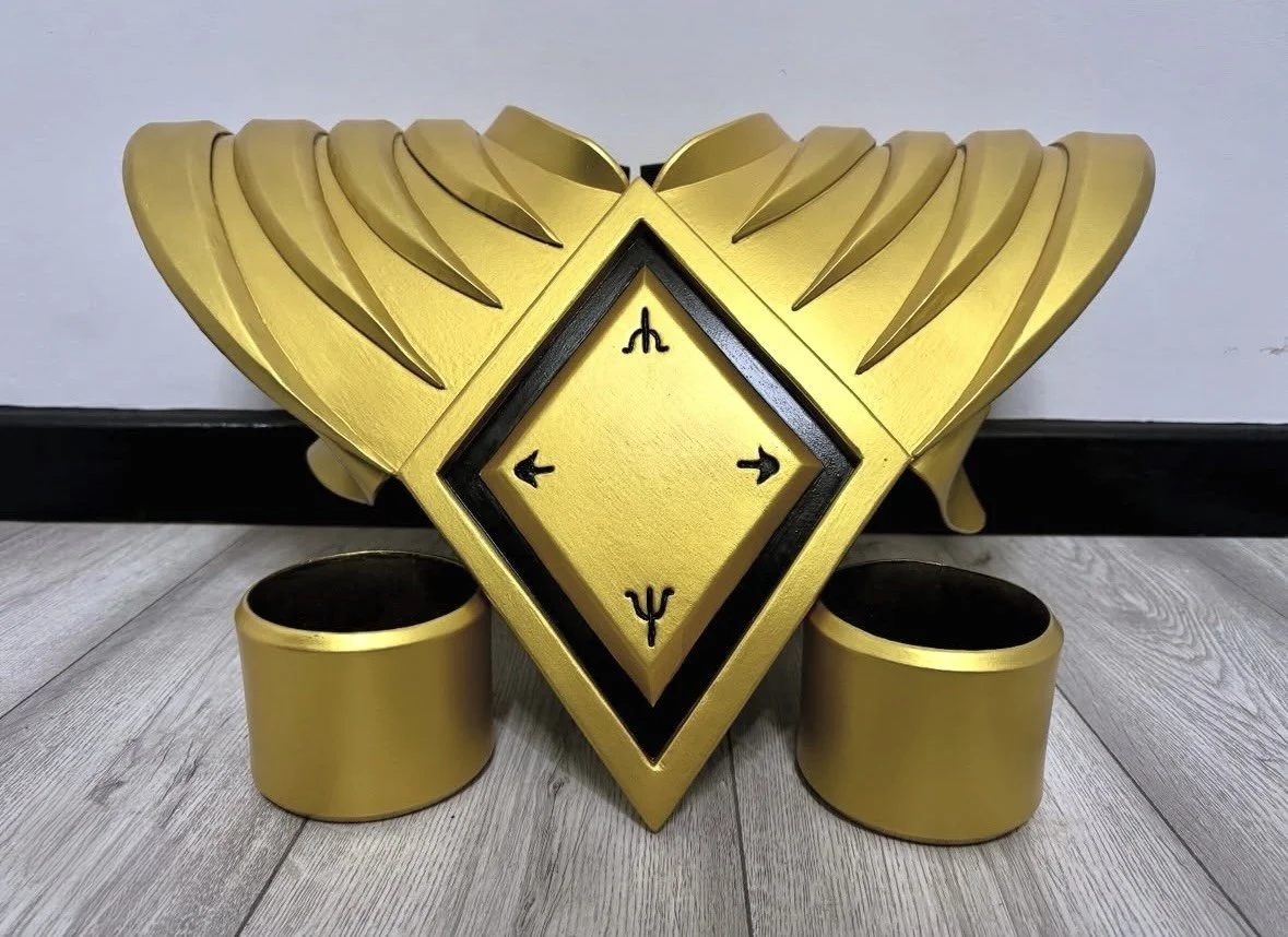 A decorative gold and black shield-shaped object with a diamond-shaped center and spiral patterns, positioned on a wooden floor, with two circular gold rings at its base.